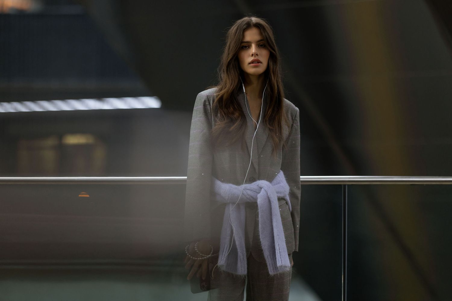 Fashion model in grey coat with white belt poses against modern interior railing with dramatic lighting.