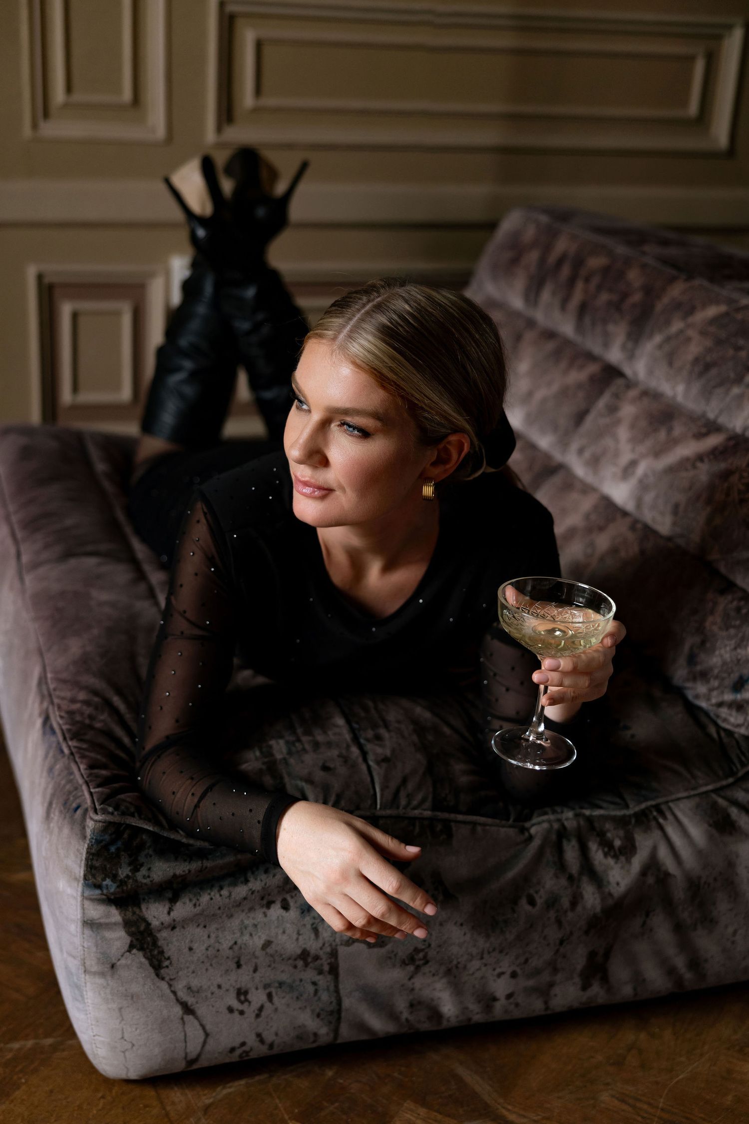 Relaxed person in black attire lounges on velvet couch with cocktail glass in moody lighting.