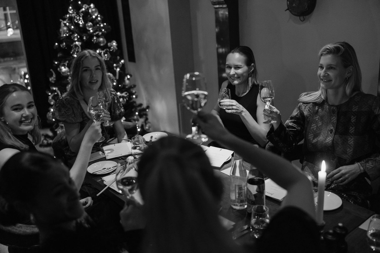 Group toasting with glasses around candlelit dinner table with Christmas tree in background.