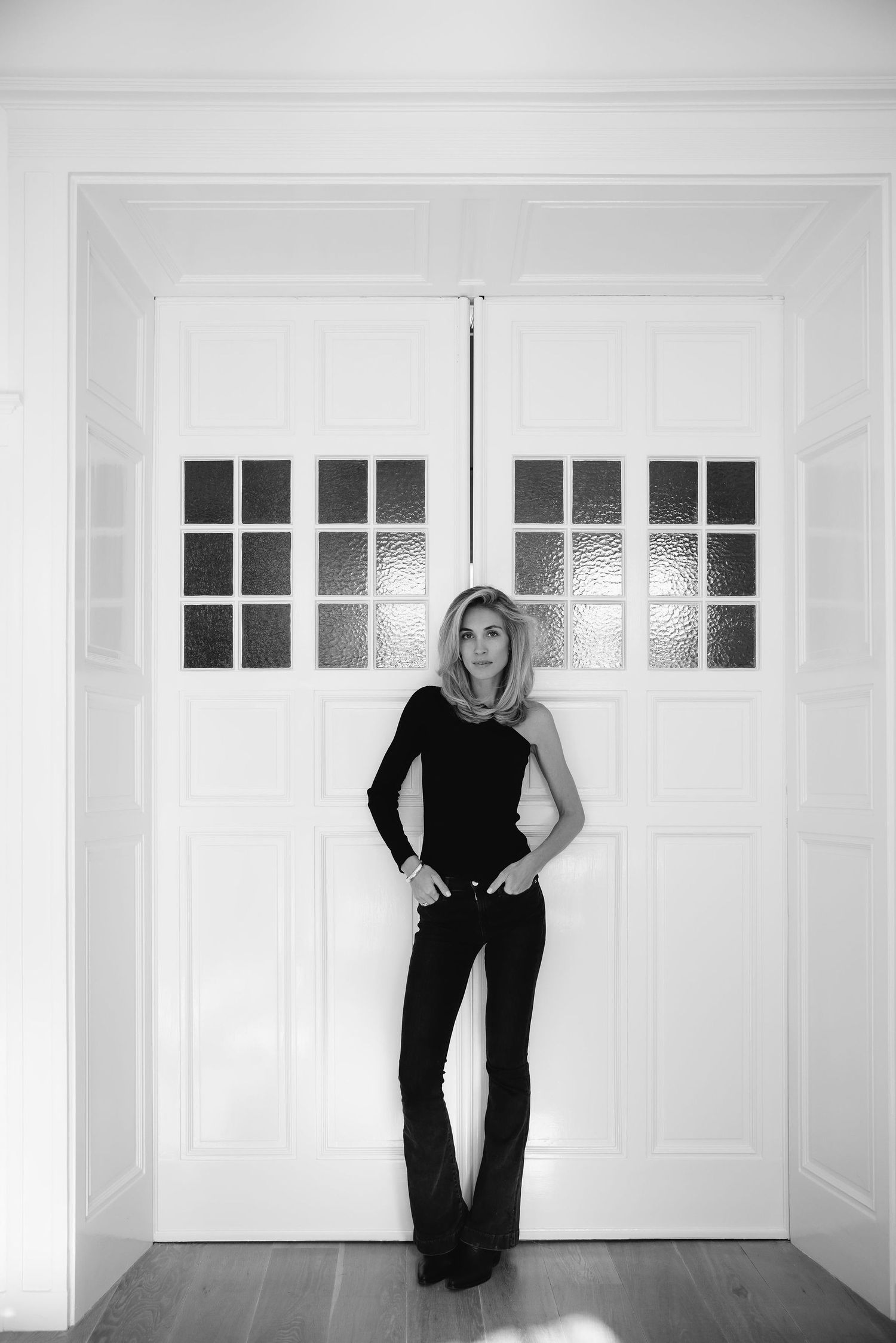 Black and white full length portrait against white double doors in minimalist setting.