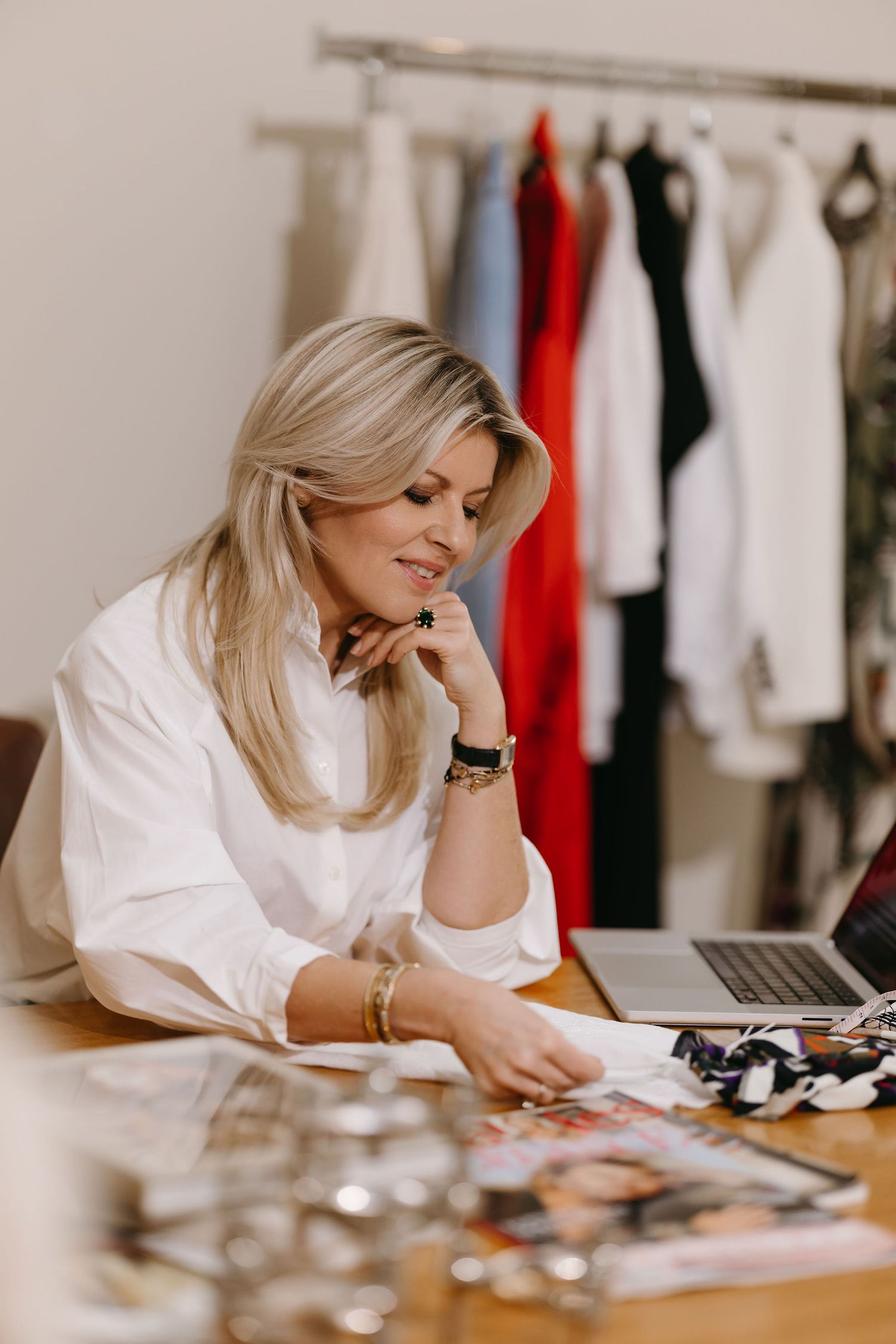 Creative professional reviewing fashion materials and accessories at a desk with clothing rack in background.