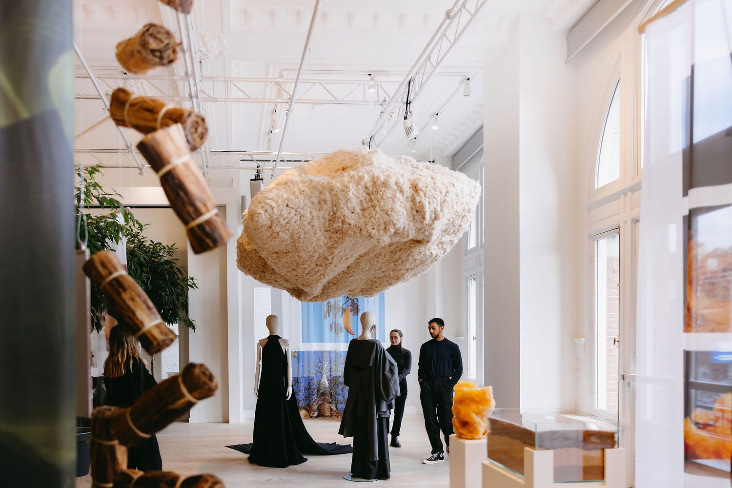 Art gallery installation featuring suspended wooden sculptures and clothing displays in bright interior space.