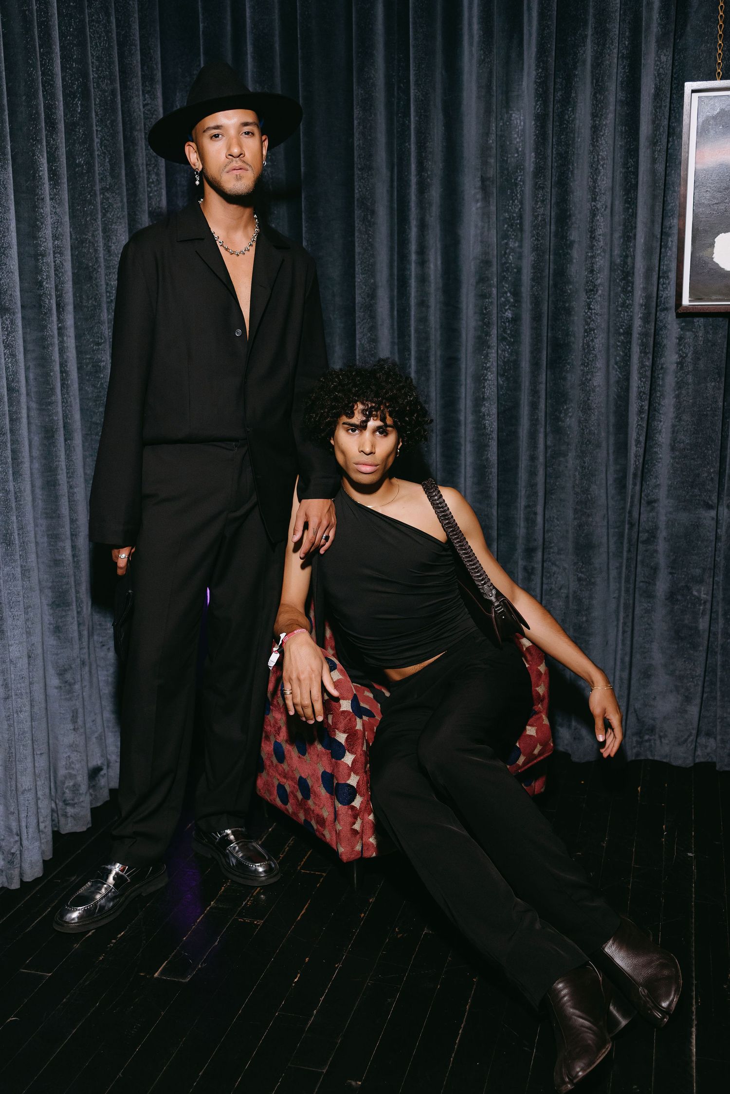 Two people in black outfits striking dramatic poses against dark curtain backdrop.