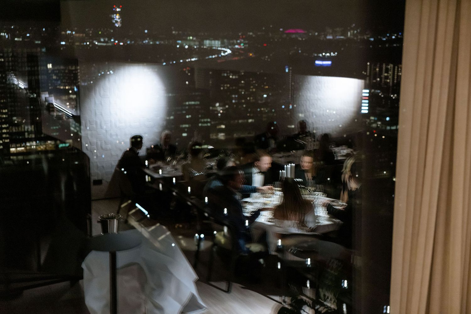A moody nighttime cityscape view through large windows with silhouettes and candlelight.