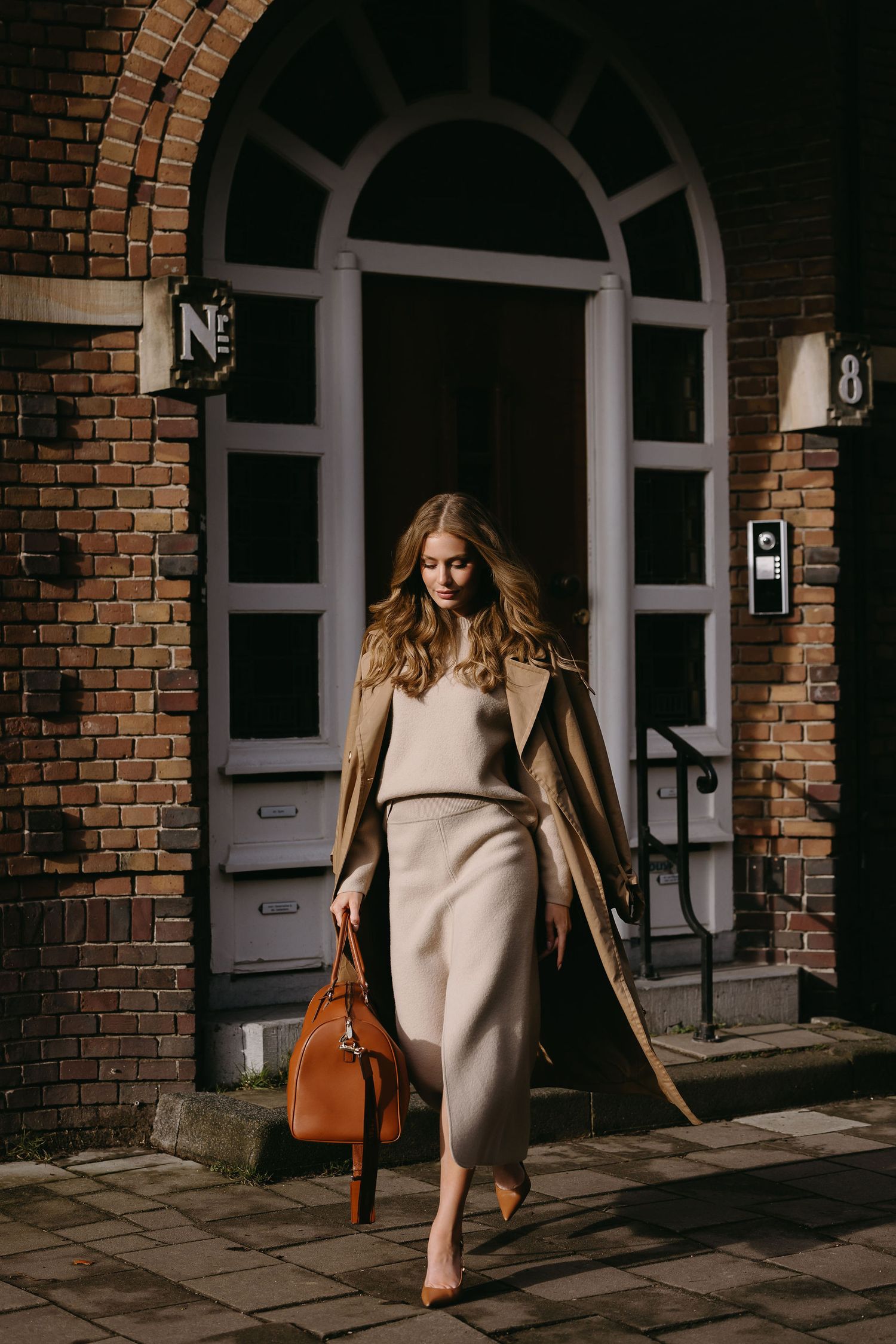 Elegant woman in beige dress and coat carrying brown leather bag outside brick building.
