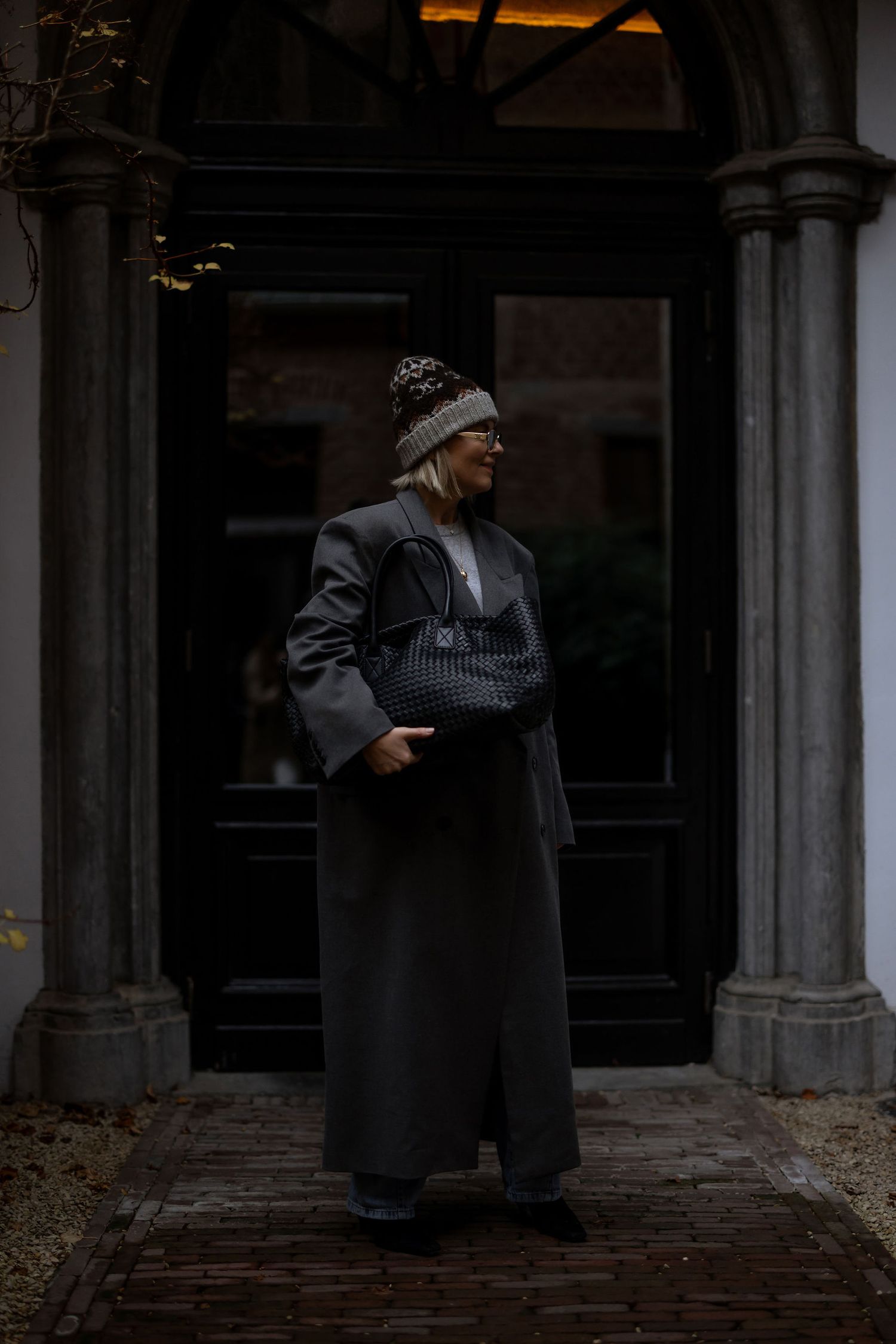 Dark moody portrait in doorway wearing long coat and knit beanie against architectural columns.