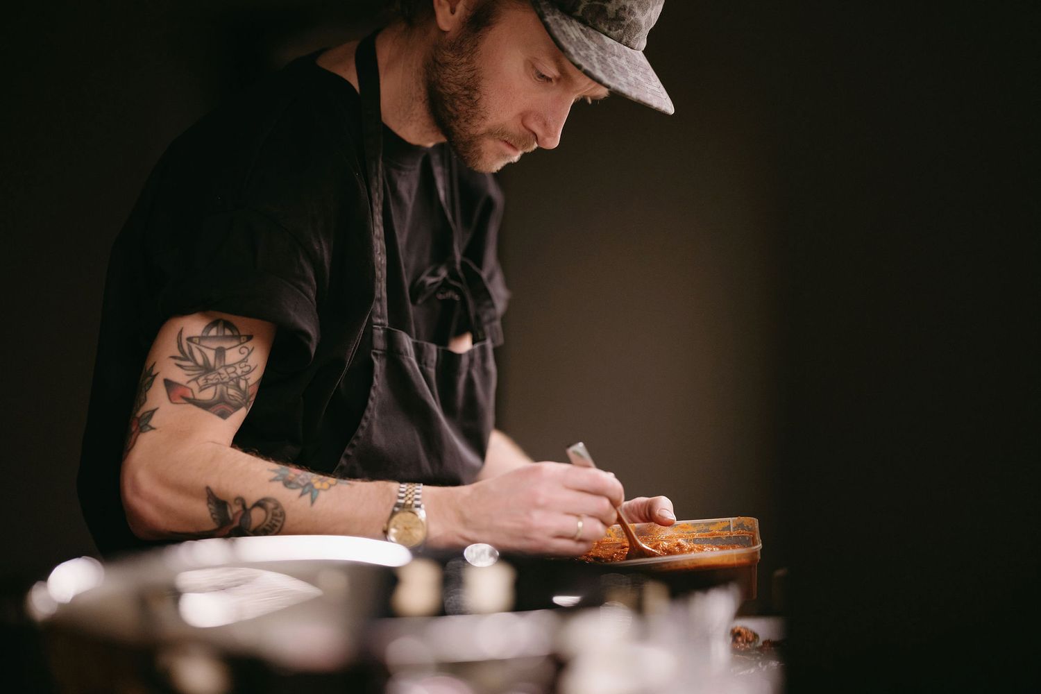 A chef with tattoos and a cap carefully prepares food at a wooden cutting board.