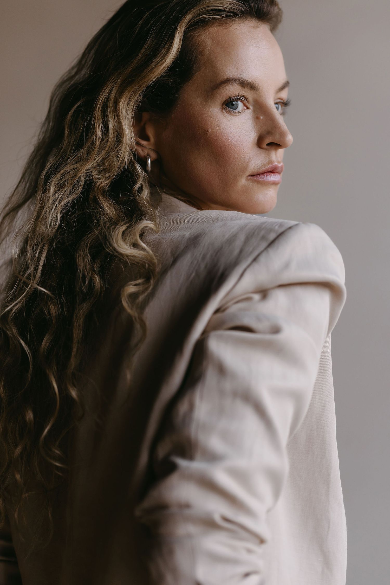 Side profile fashion portrait showing wavy blonde hair and a beige blazer against a neutral background.