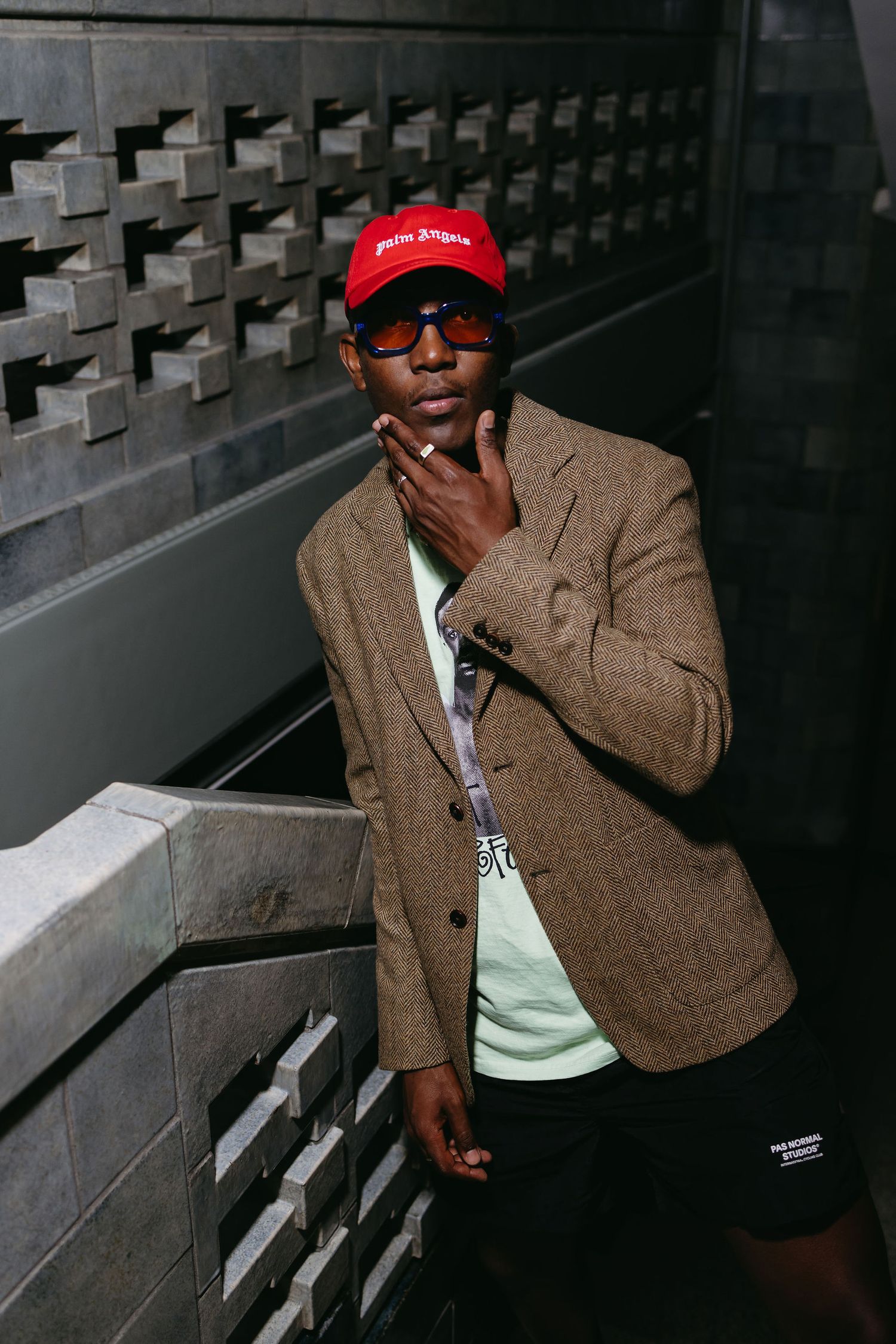 Stylish outfit with brown tweed blazer, red cap and sunglasses against industrial metal wall background.
