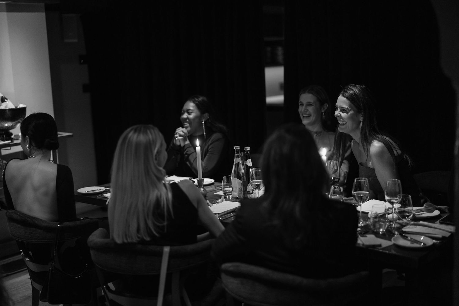 Black and white photo of people gathered around dinner table in intimate setting.
