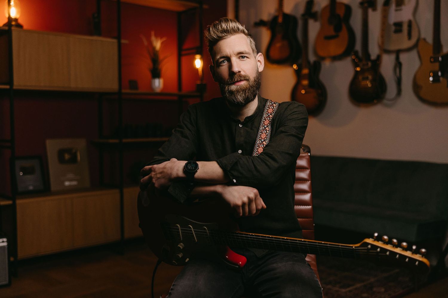 Musician in dark outfit sitting in music studio with guitars on wall.