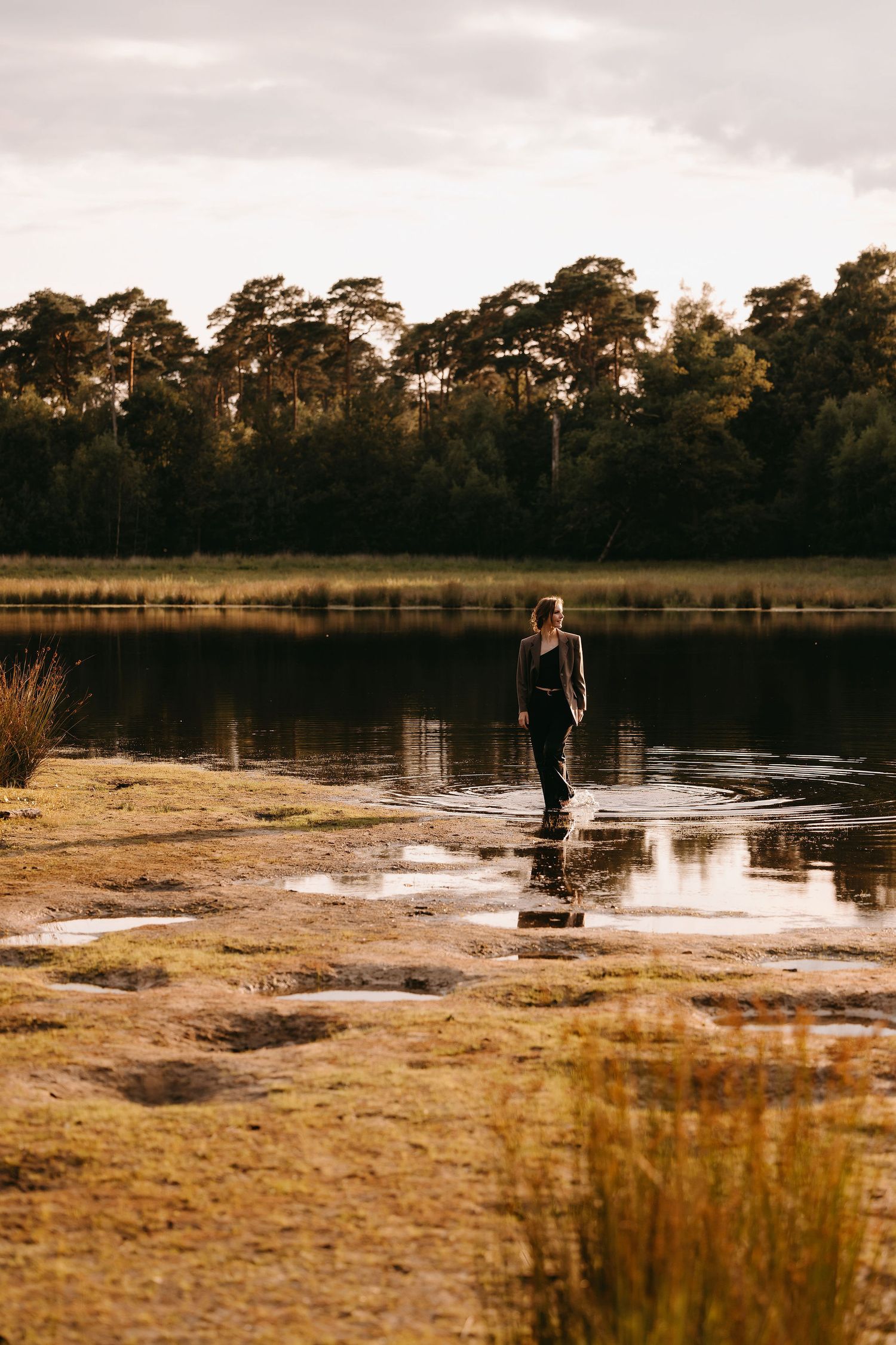 Atmospheric landscape photo of person standing by lake shore at sunset with pine trees.