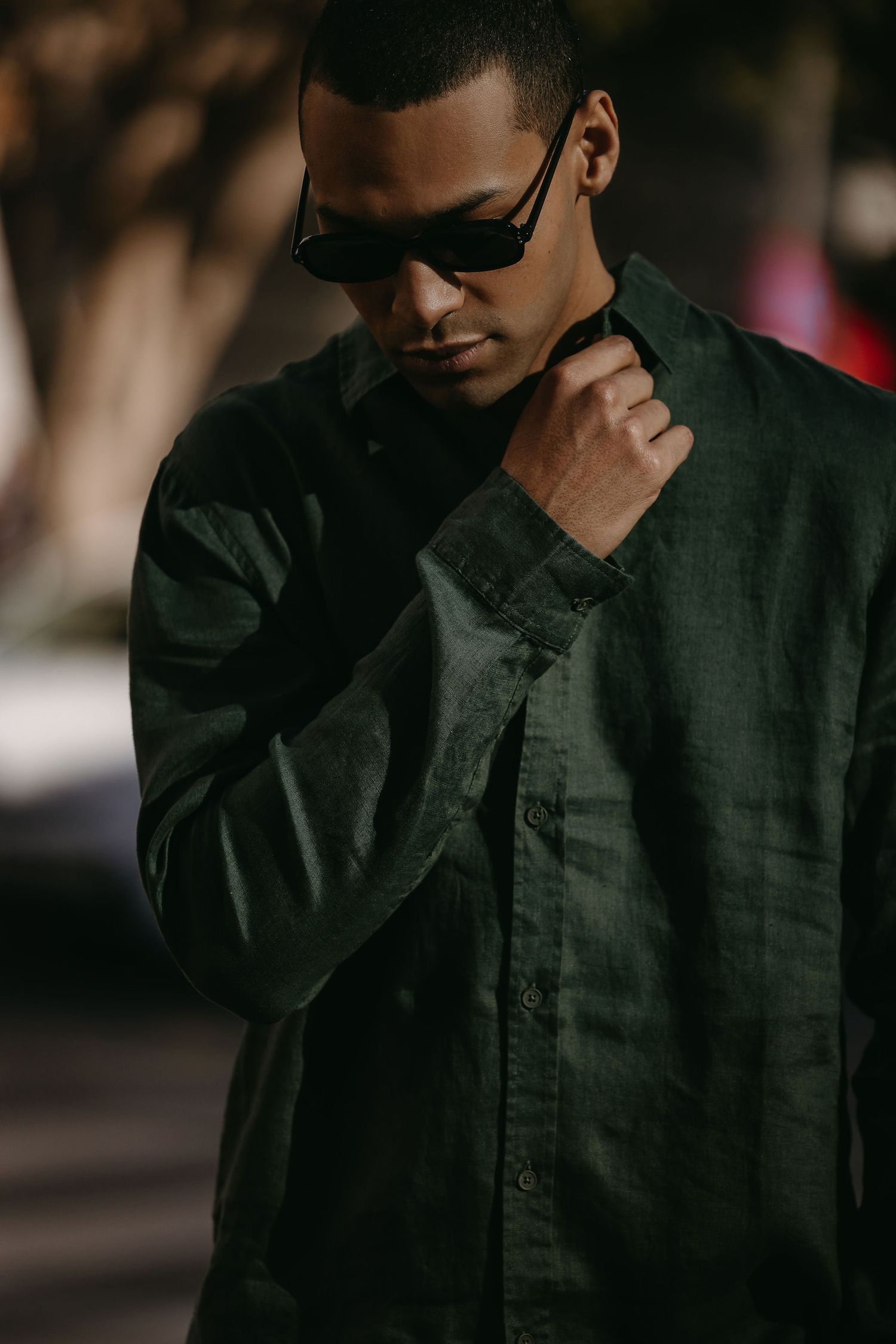 Moody portrait of someone wearing sunglasses and a dark green button-up shirt in natural lighting.