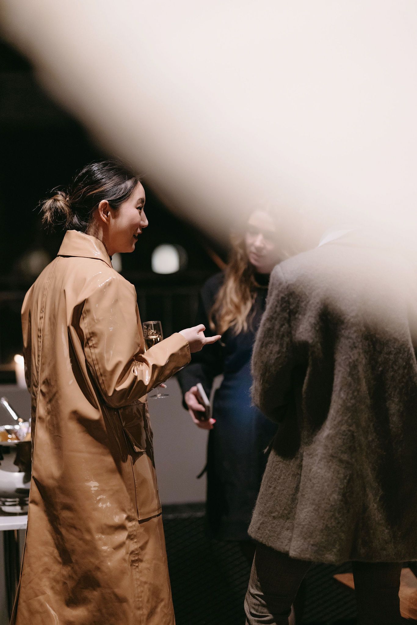 A person in a tan coat stands among attendees at an indoor gathering.