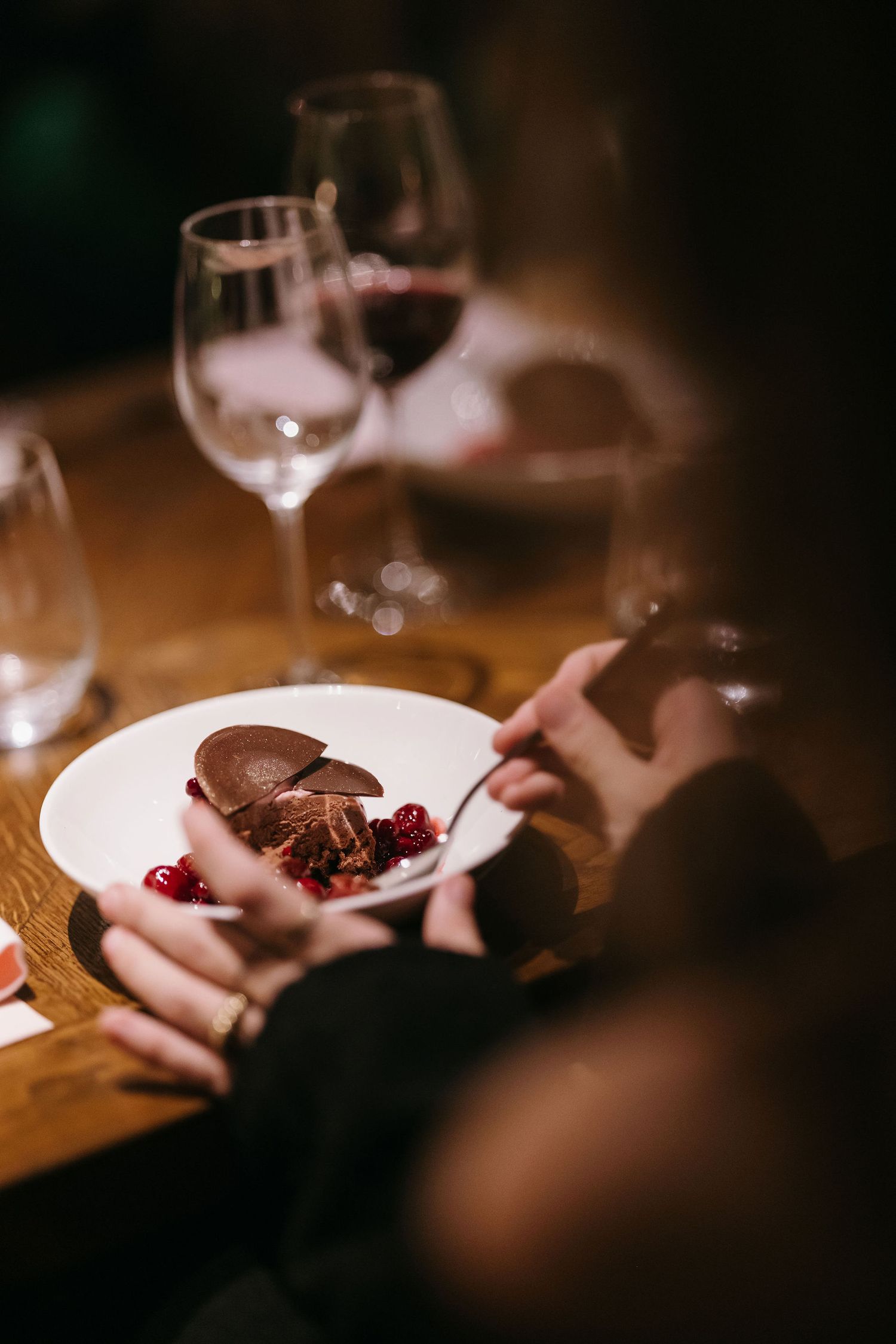 Close-up of hands holding fork over chocolate dessert with wine glasses in background.