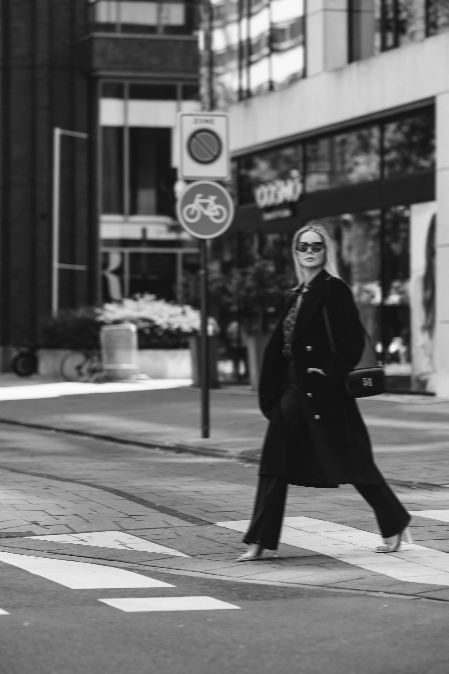 Black and white urban street photo showing pedestrian in long coat crossing at bicycle sign.