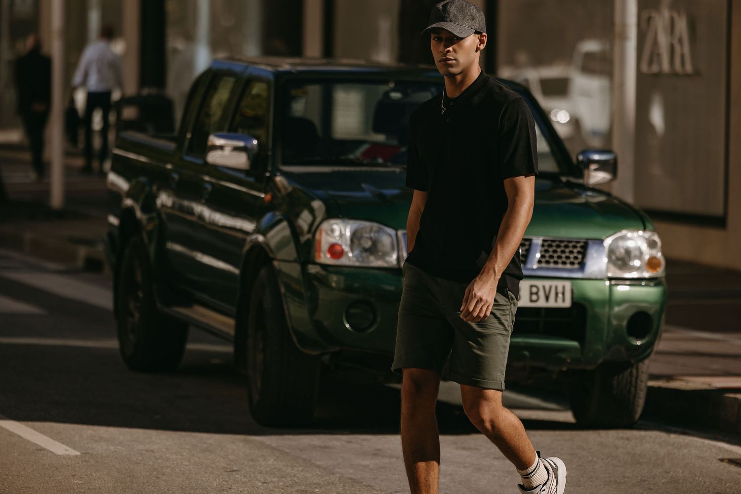 Street style shot of person in casual black outfit walking past parked car at dusk.