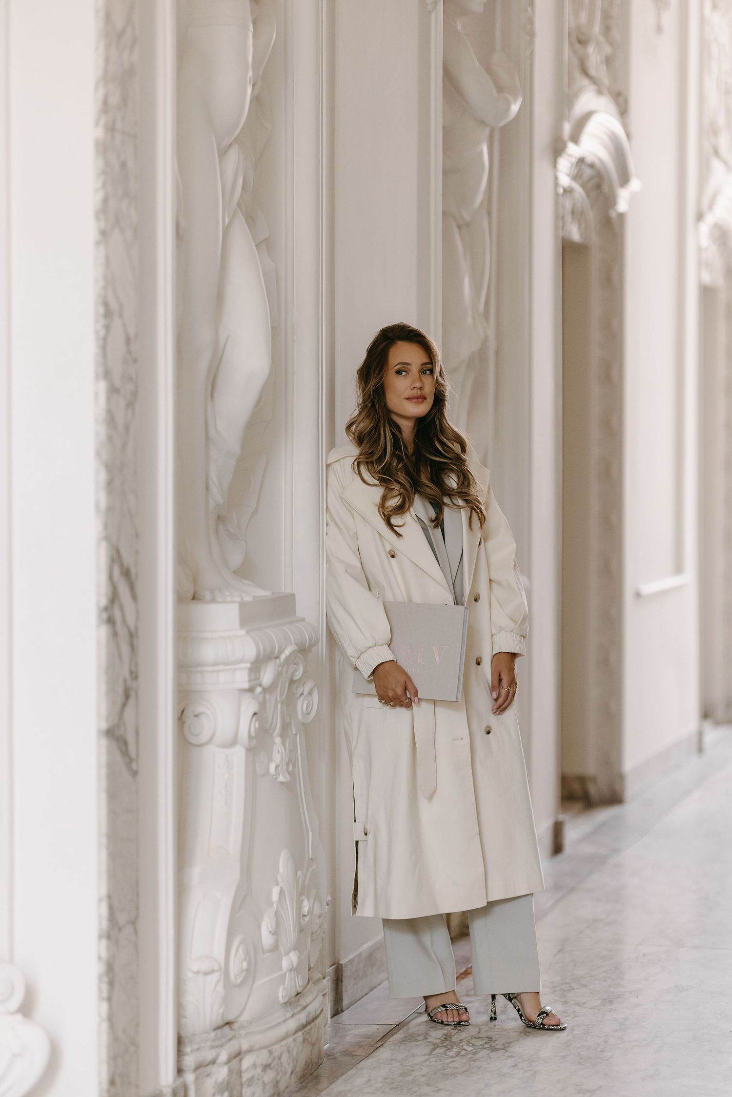 Elegant white trench coat look photographed in ornate classical hallway with decorative moldings.