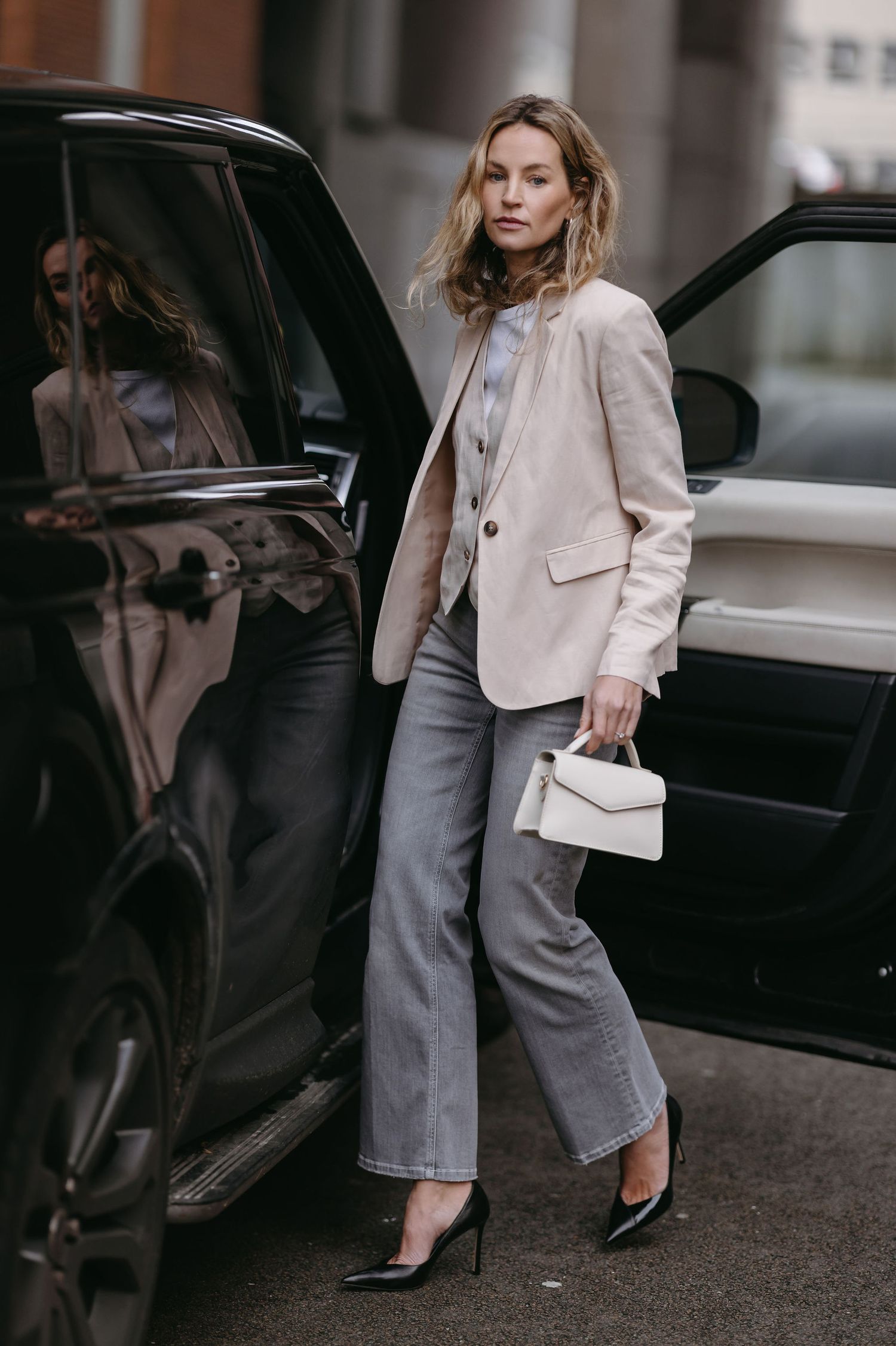 Street style shot of someone exiting a black car wearing a beige blazer and gray trousers.