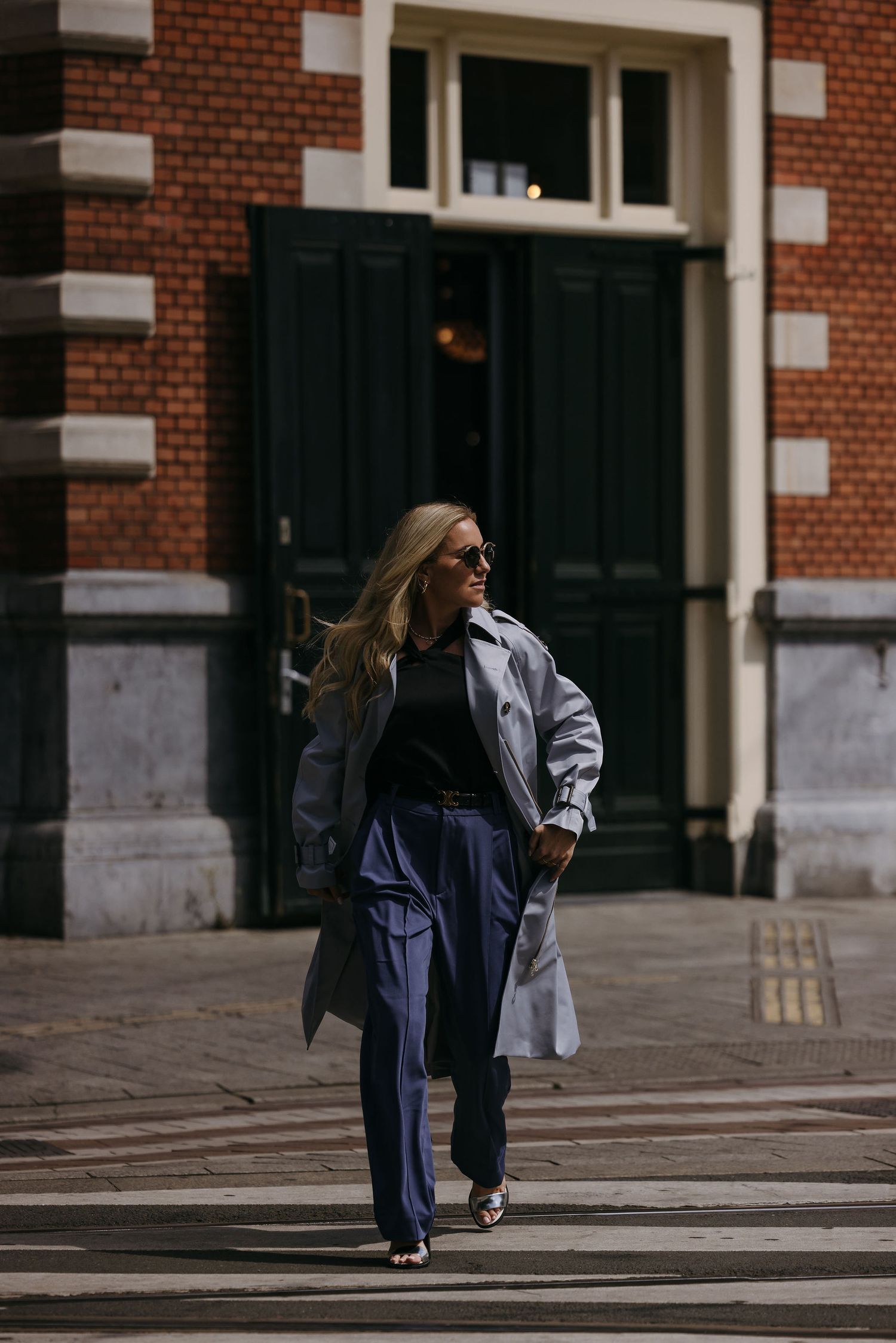 Street style fashion shot featuring long grey coat and wide-leg pants against brick building backdrop.