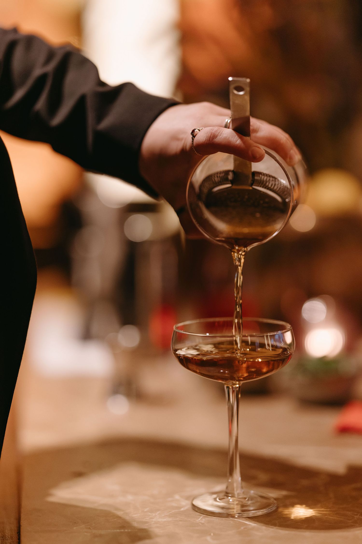 A close-up of someone pouring a cocktail into a coupe glass at a bar.
