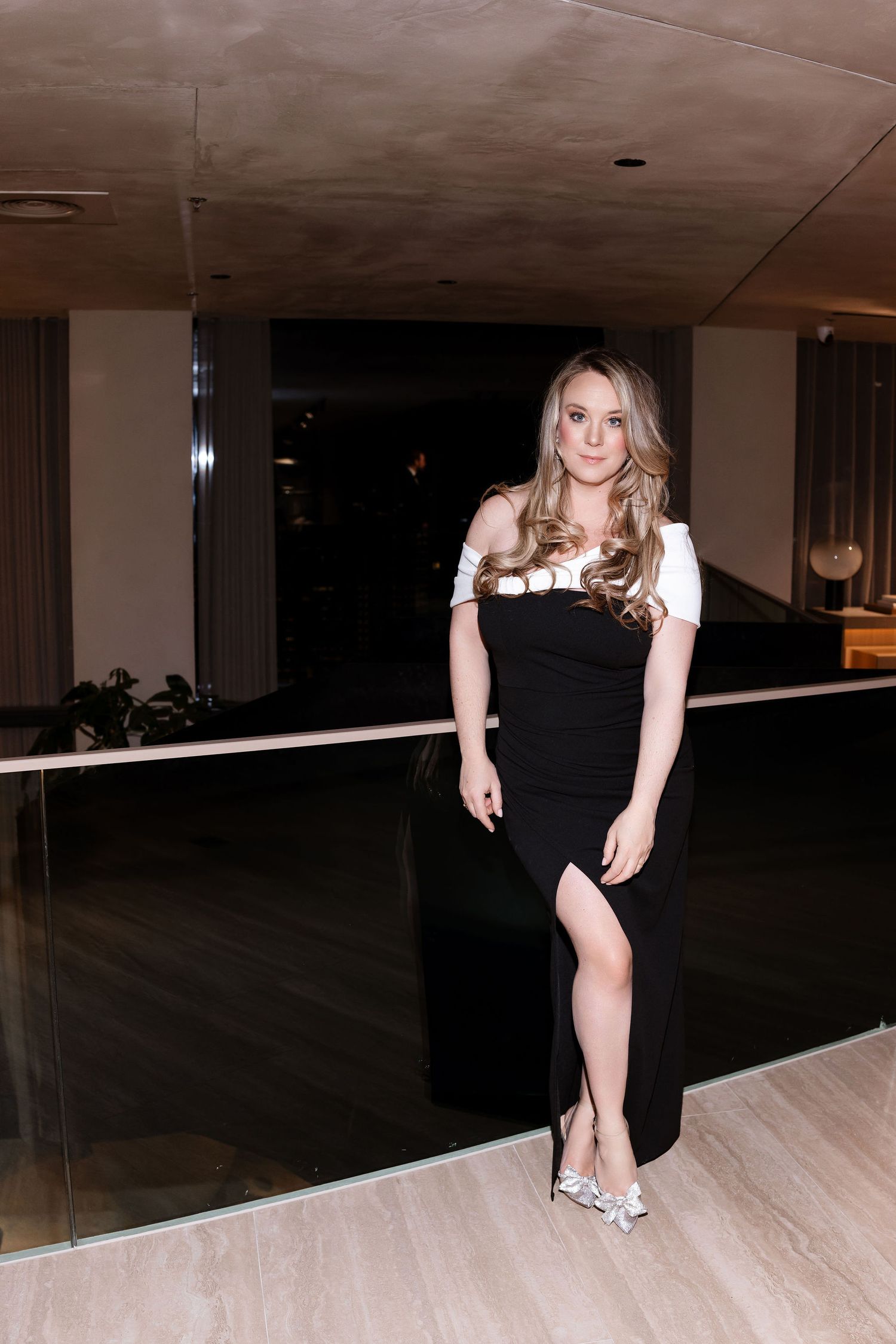 Woman in black and white off-shoulder dress poses in modern interior setting.