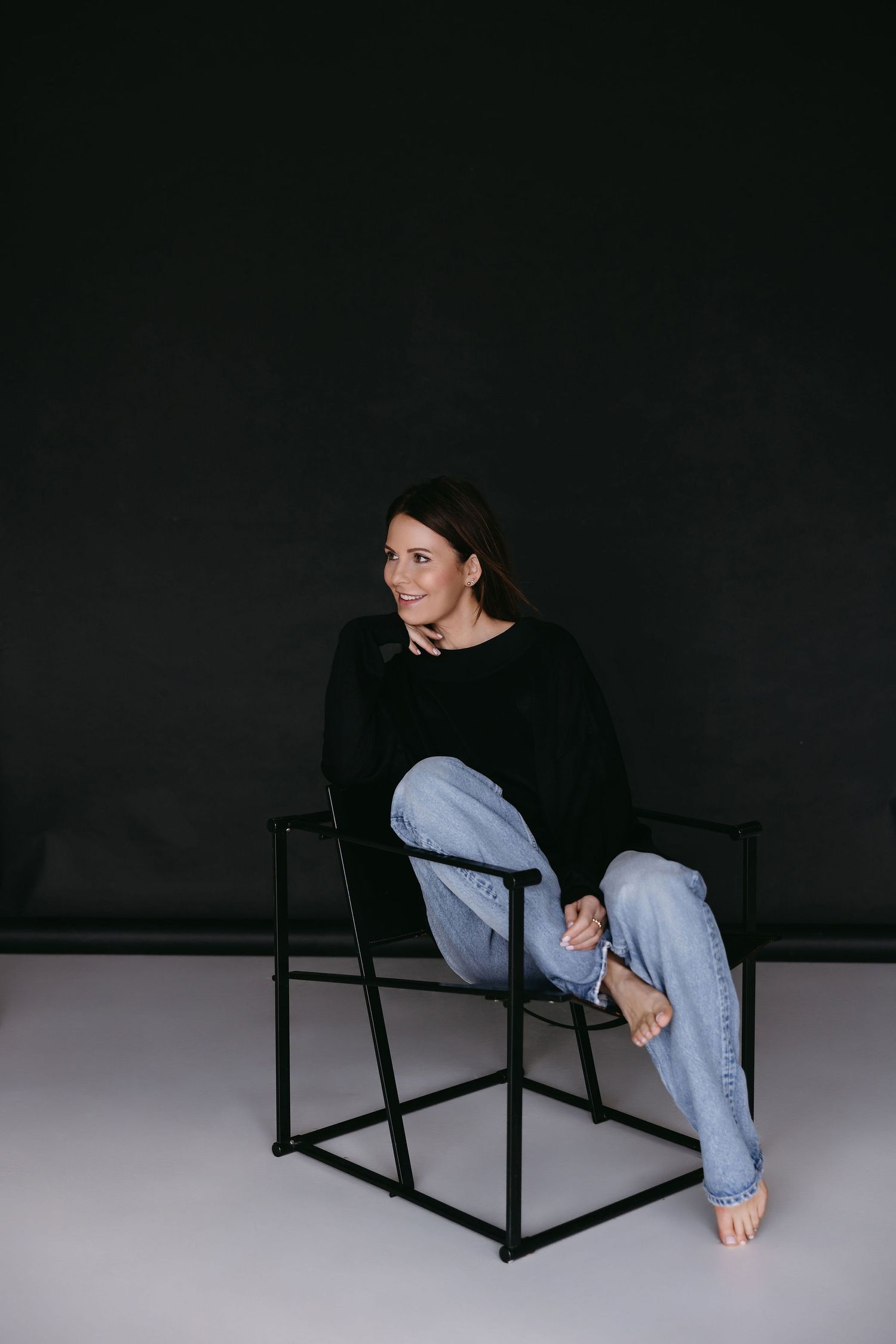 Casual portrait of person sitting in minimalist black chair against dark background.