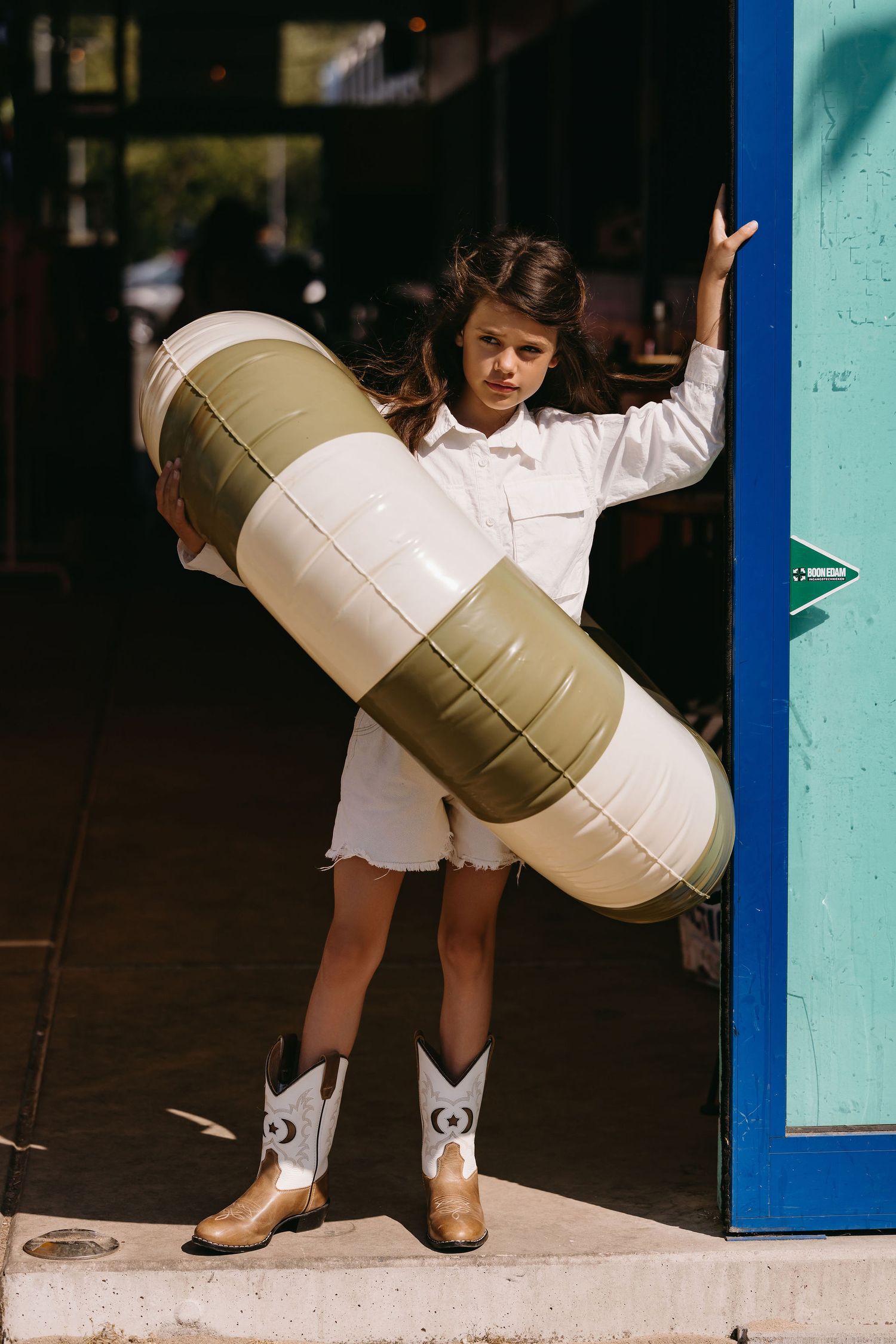 Young person in white outfit holds inflatable pool float while wearing cowboy boots.