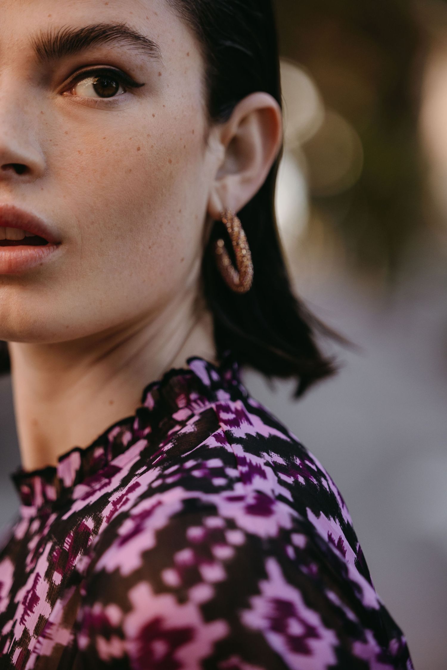 Close-up fashion portrait featuring purple patterned top and textured hoop earrings against blurred background.