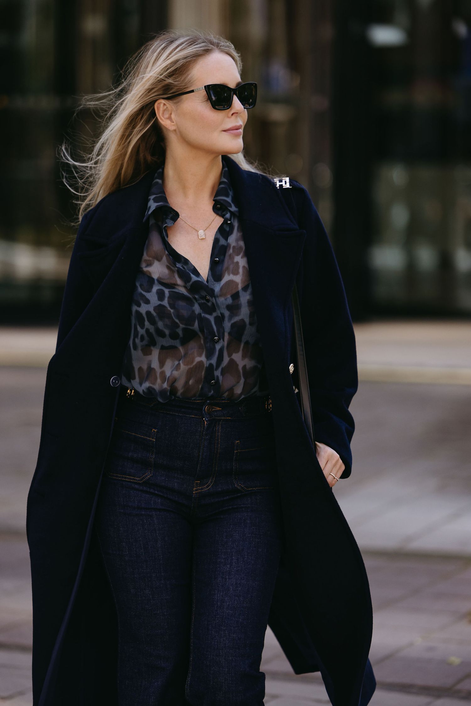 Stylish woman in black coat and leopard print blouse walking outdoors wearing sunglasses.