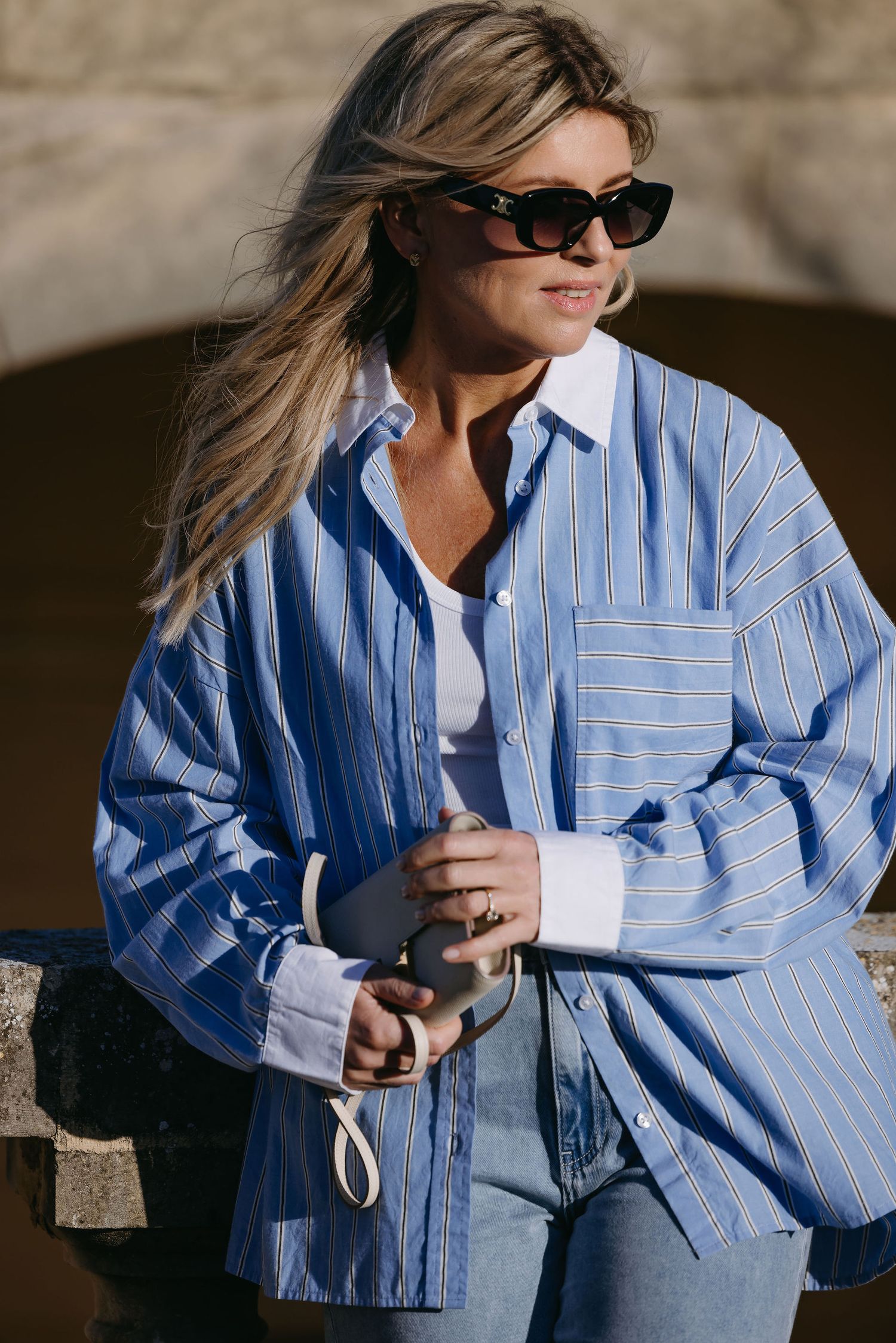 Stylish woman in oversized blue striped shirt and sunglasses poses in natural sunlight.