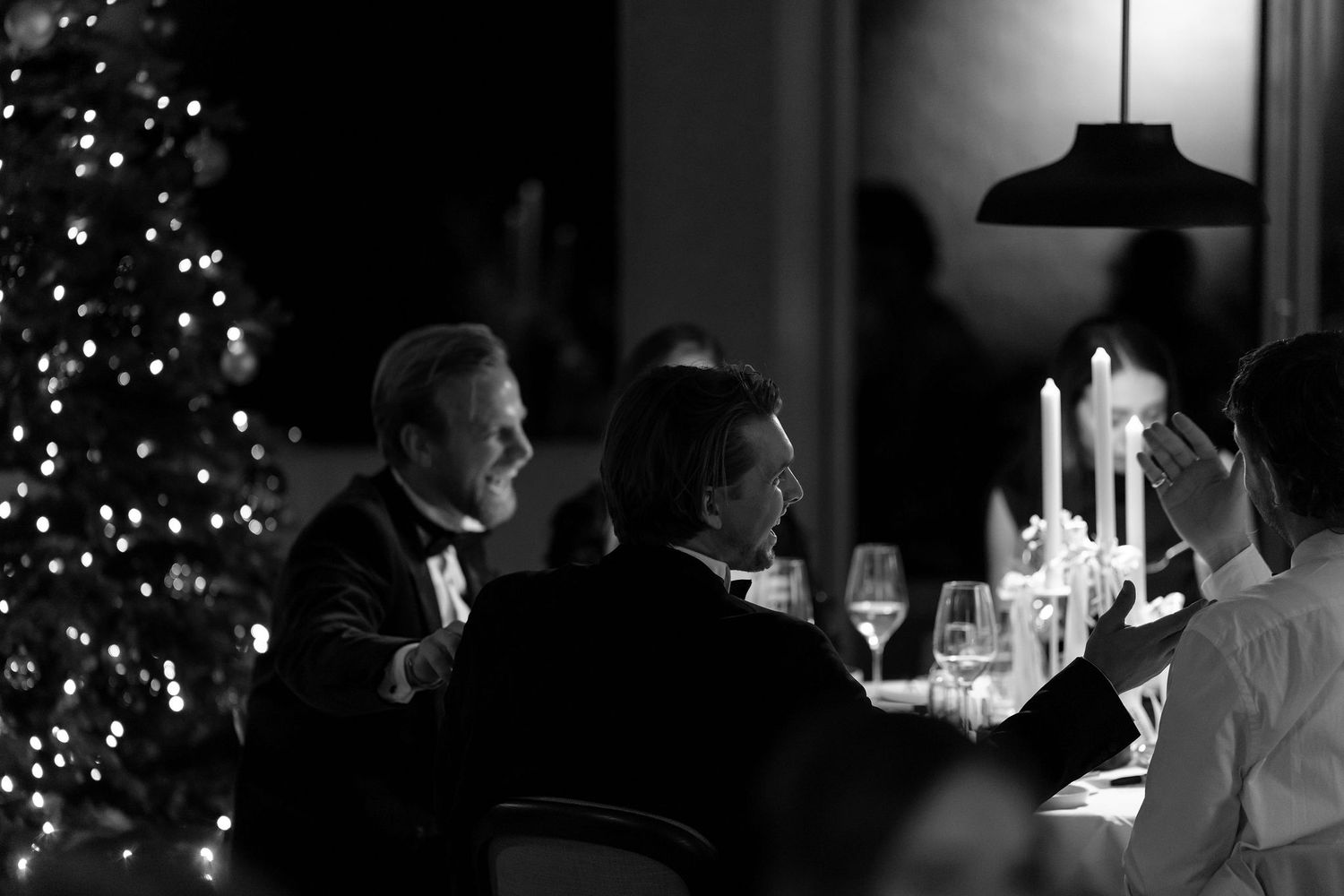 A black and white photo of an elegant dinner party with a Christmas tree glowing in the background.