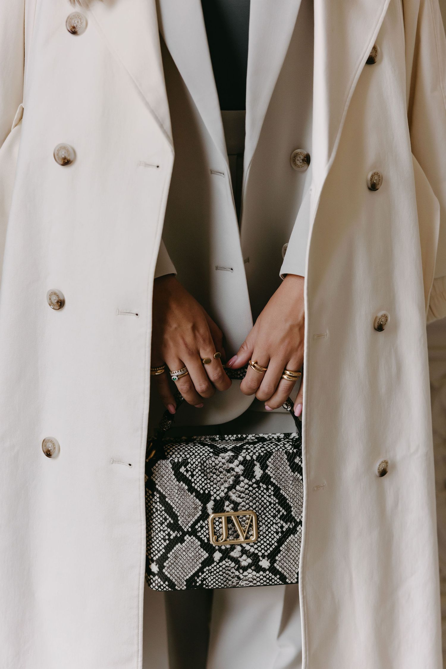 Fashion detail shot of cream coat and snakeskin purse with jewelry-adorned hands.