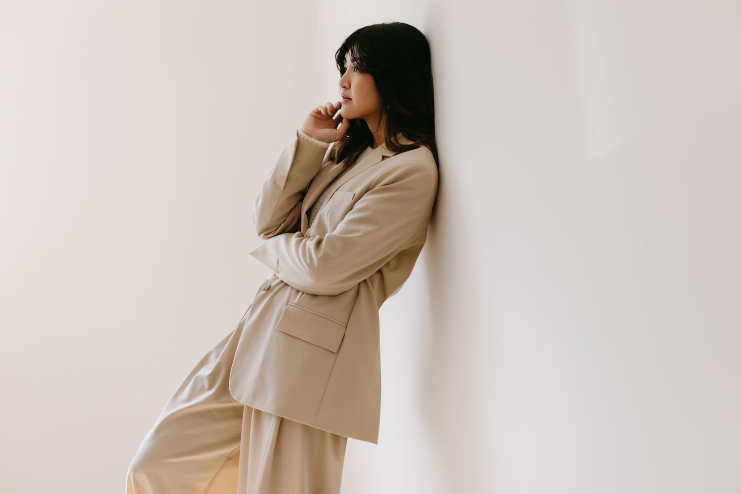 Woman in beige business suit poses thoughtfully against white wall in minimalist setting.