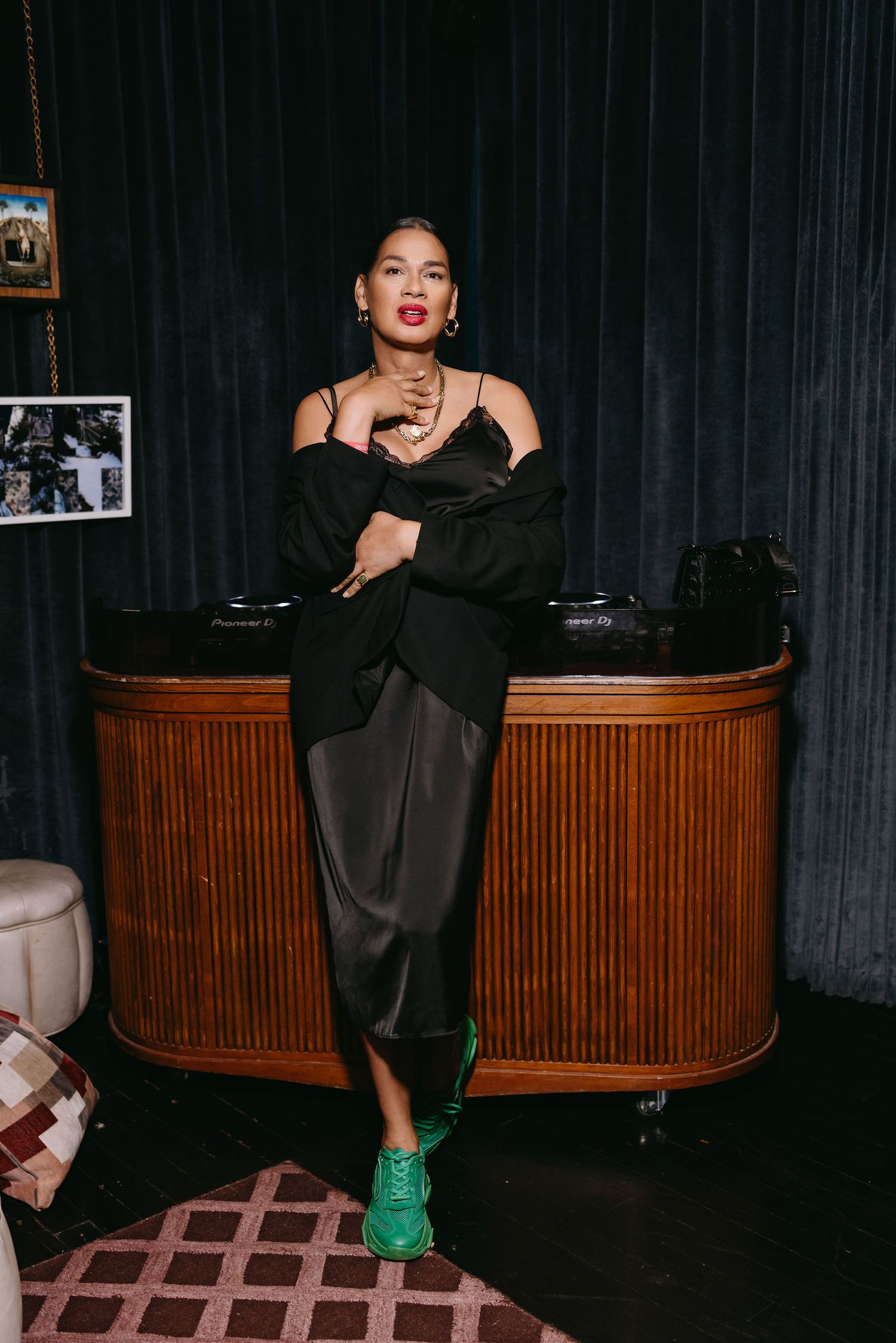 Stylish figure in black outfit and green sneakers posing against dark backdrop with vintage furniture.