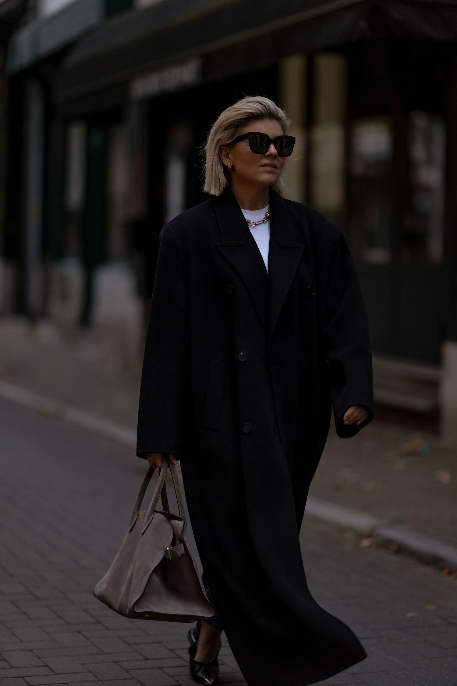 Stylish street fashion look featuring a long black coat, white top and beige handbag on a city sidewalk.