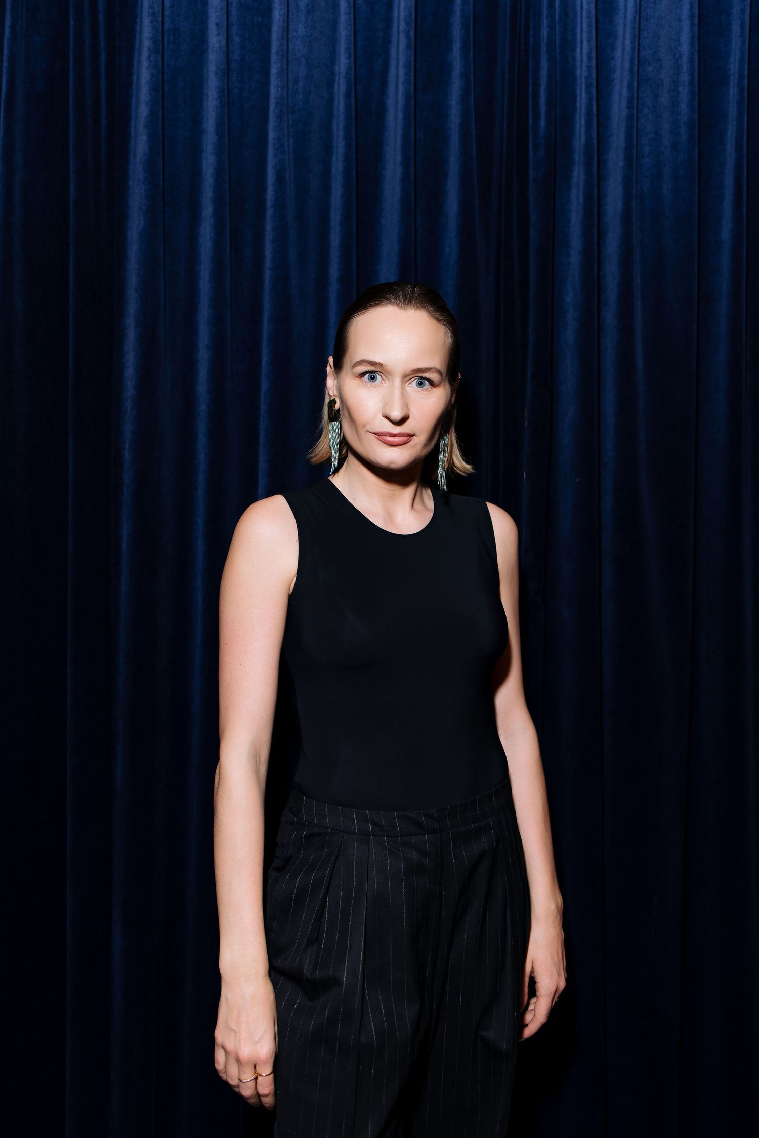 Person in black sleeveless outfit poses against dark blue curtain backdrop.