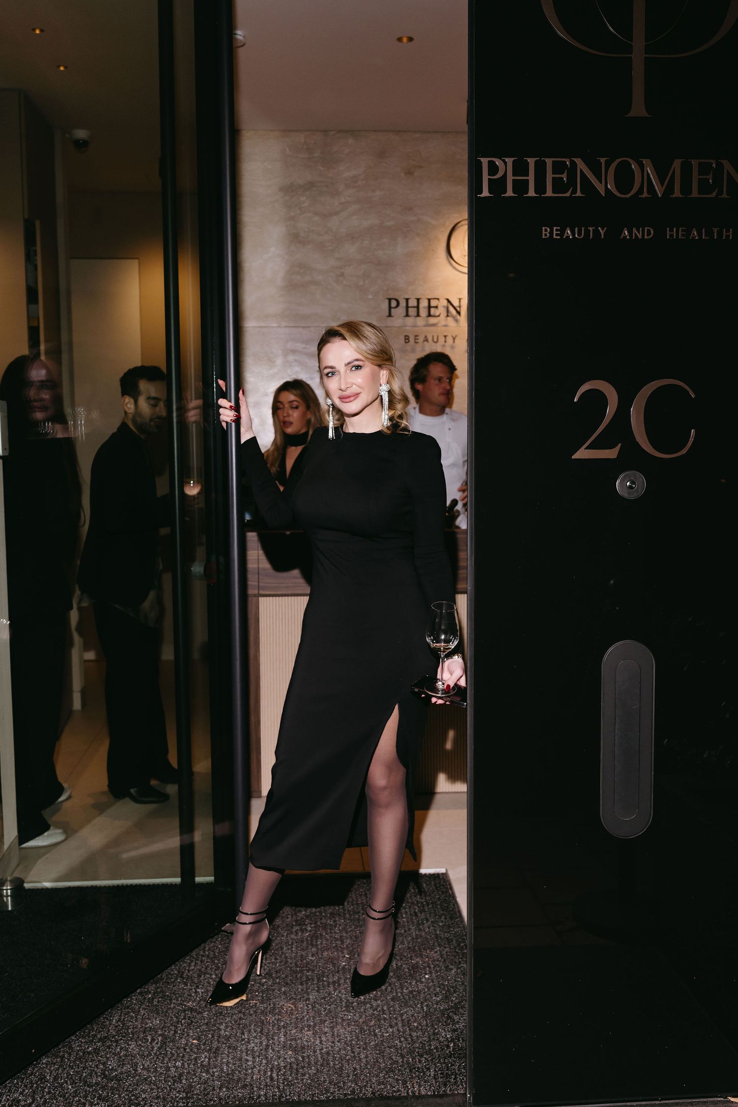 Woman in black dress entering Phenomena Beauty store doorway with wine glass.