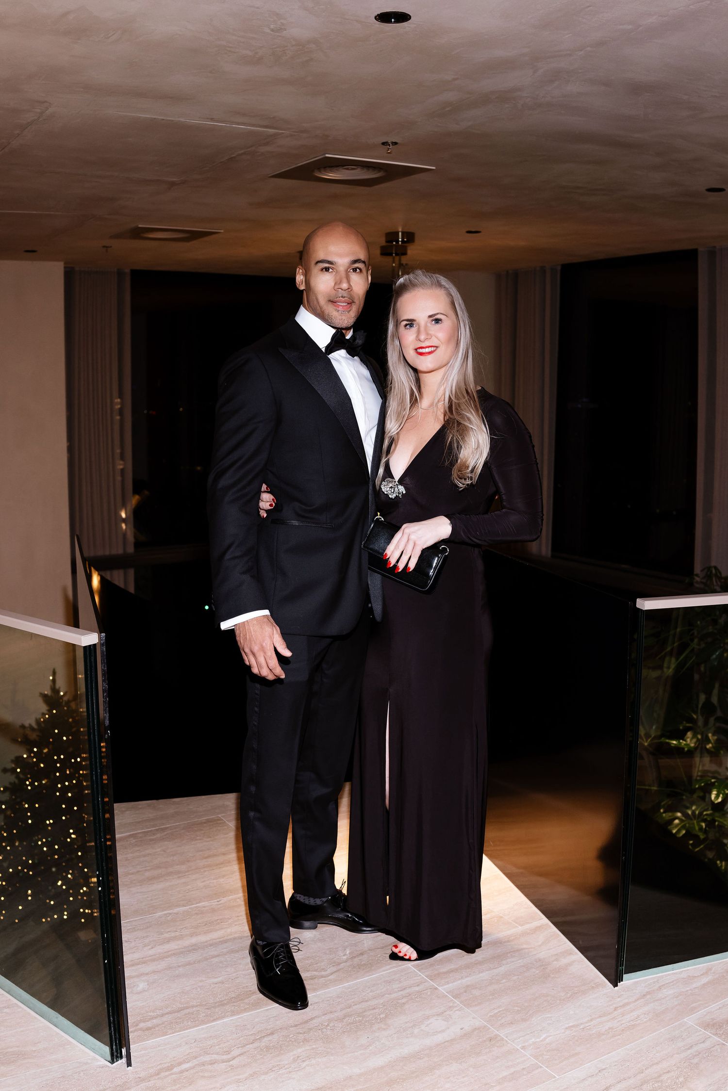 An elegant couple poses in formal black-tie attire at an evening event with a Christmas tree in the background.