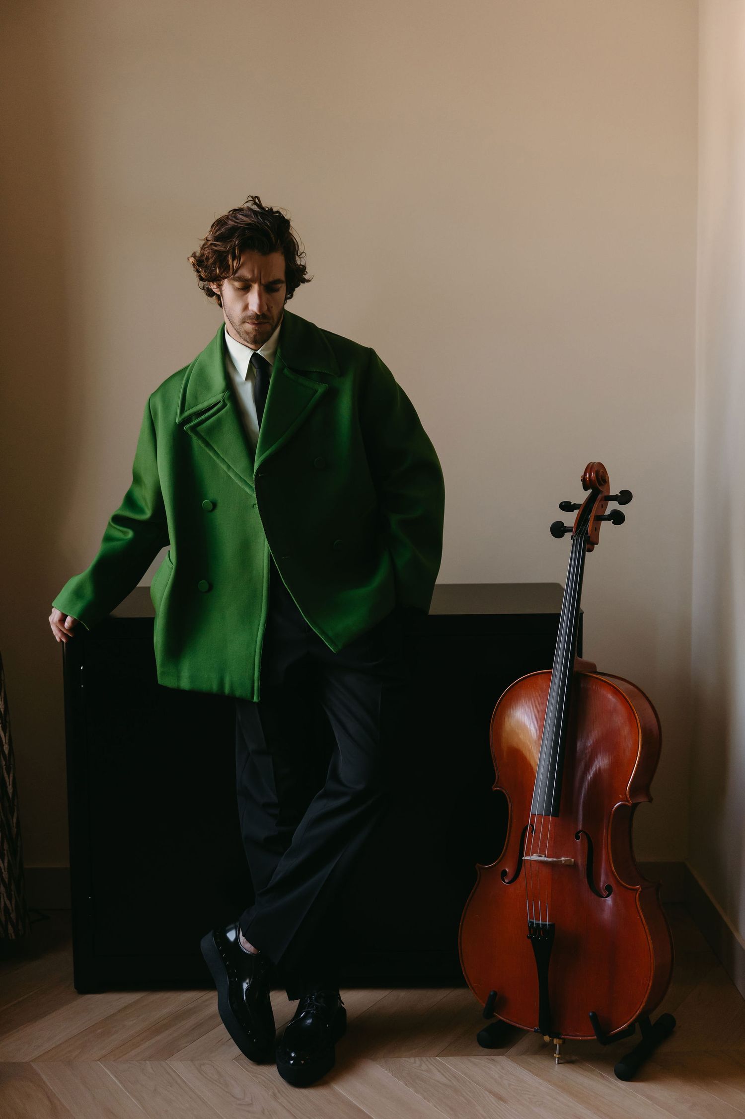 Musician in bright green coat poses next to cello against neutral background.