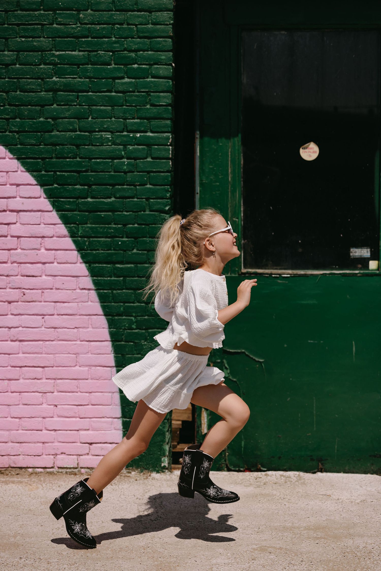 Playful scene of someone in white outfit and combat boots running past pink and green brick wall.