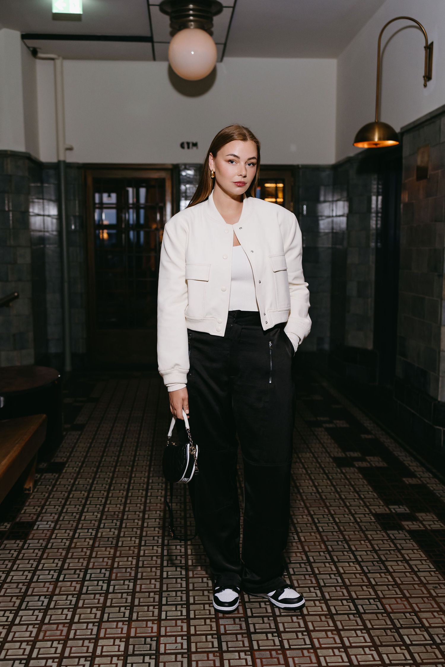Streetwear fashion look featuring white bomber jacket, black pants and sneakers in a vintage tiled hallway setting.