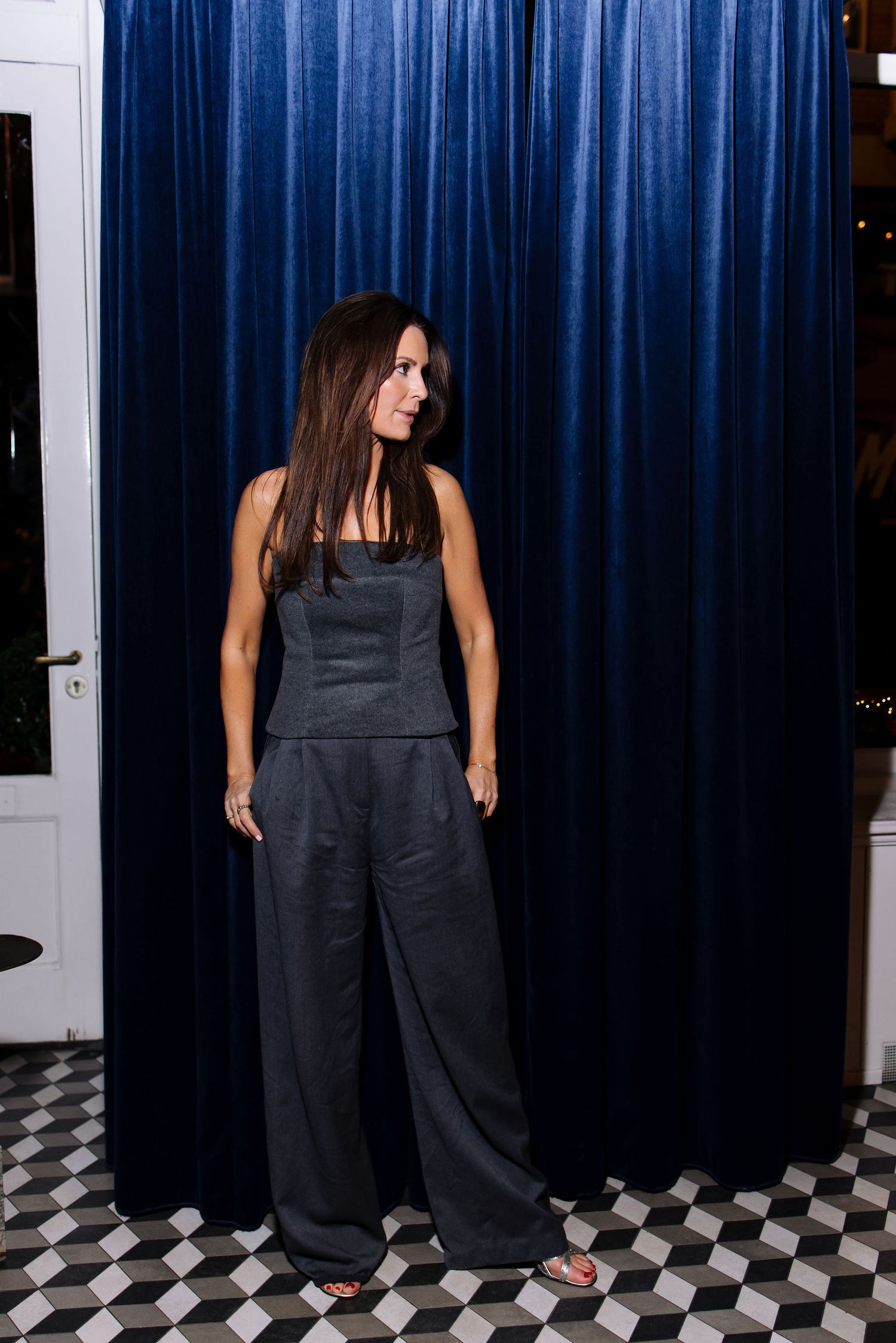 Woman in dark strapless jumpsuit poses against blue velvet curtain backdrop.