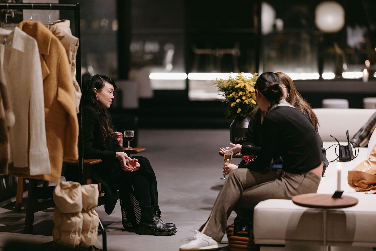 People engaged in conversation near a clothing rack in a dimly lit retail or event space.