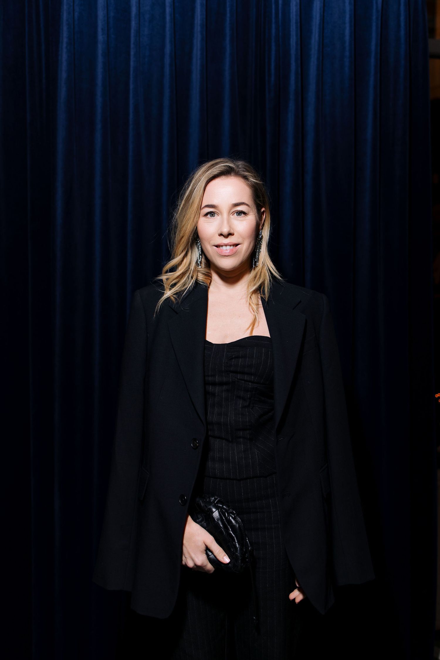 Person wearing black blazer and dress poses against blue curtain background.