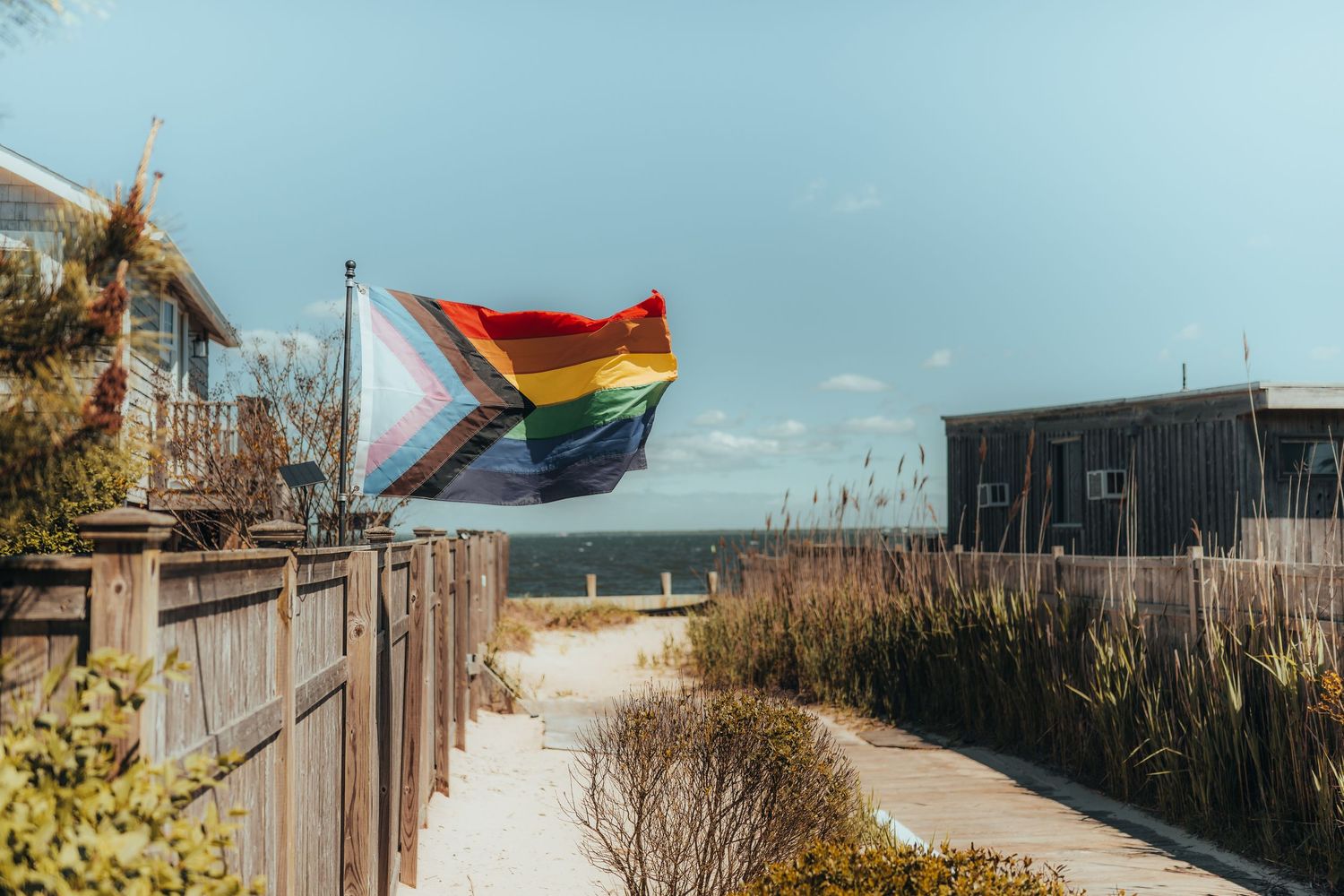 Progress Pride flag waving in the breeze along a sandy beach pathway.