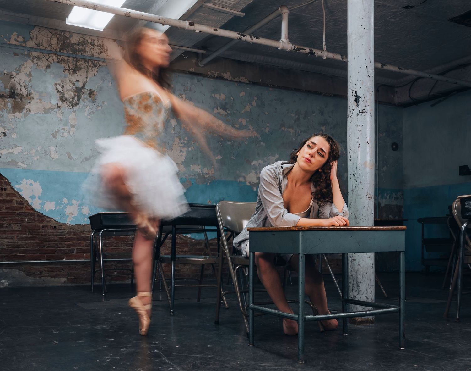 Two women in an abandoned room, one a ballerina dancing en pointe, the other seated at a desk looking pensive.