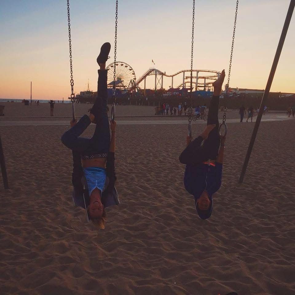 Two people hanging upside down on swings at Santa Monica beach pier at sunset.