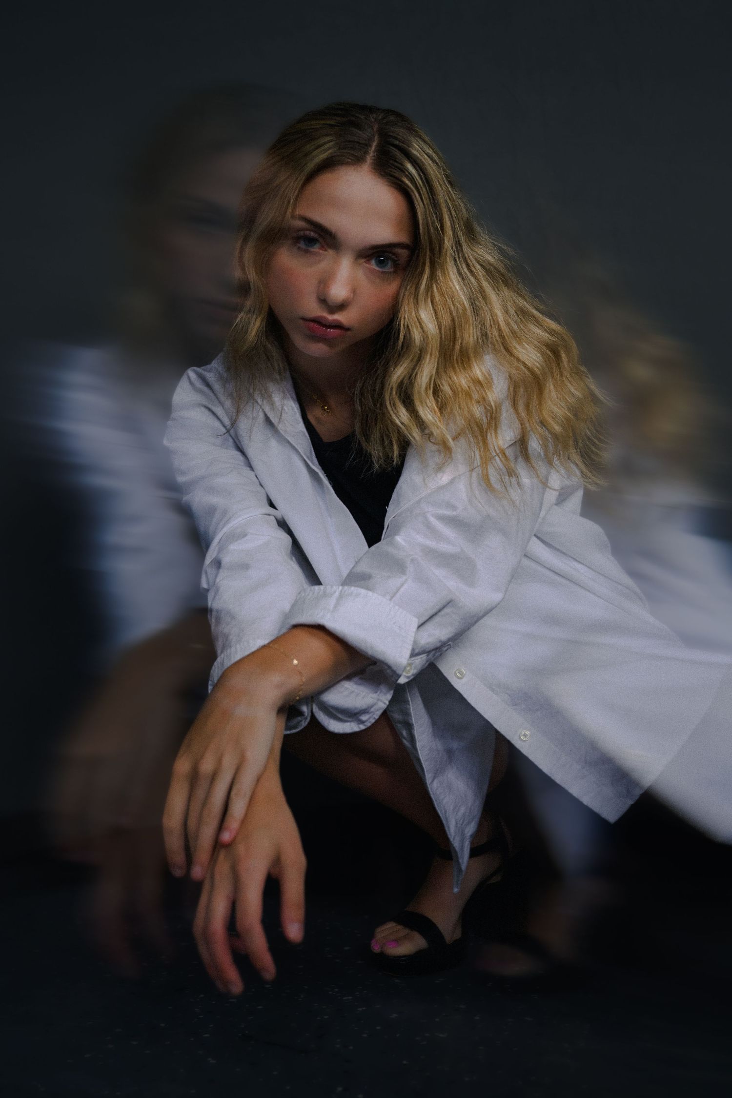 Blonde woman in white blazer sits pensively against dark background with artistic motion blur effect.