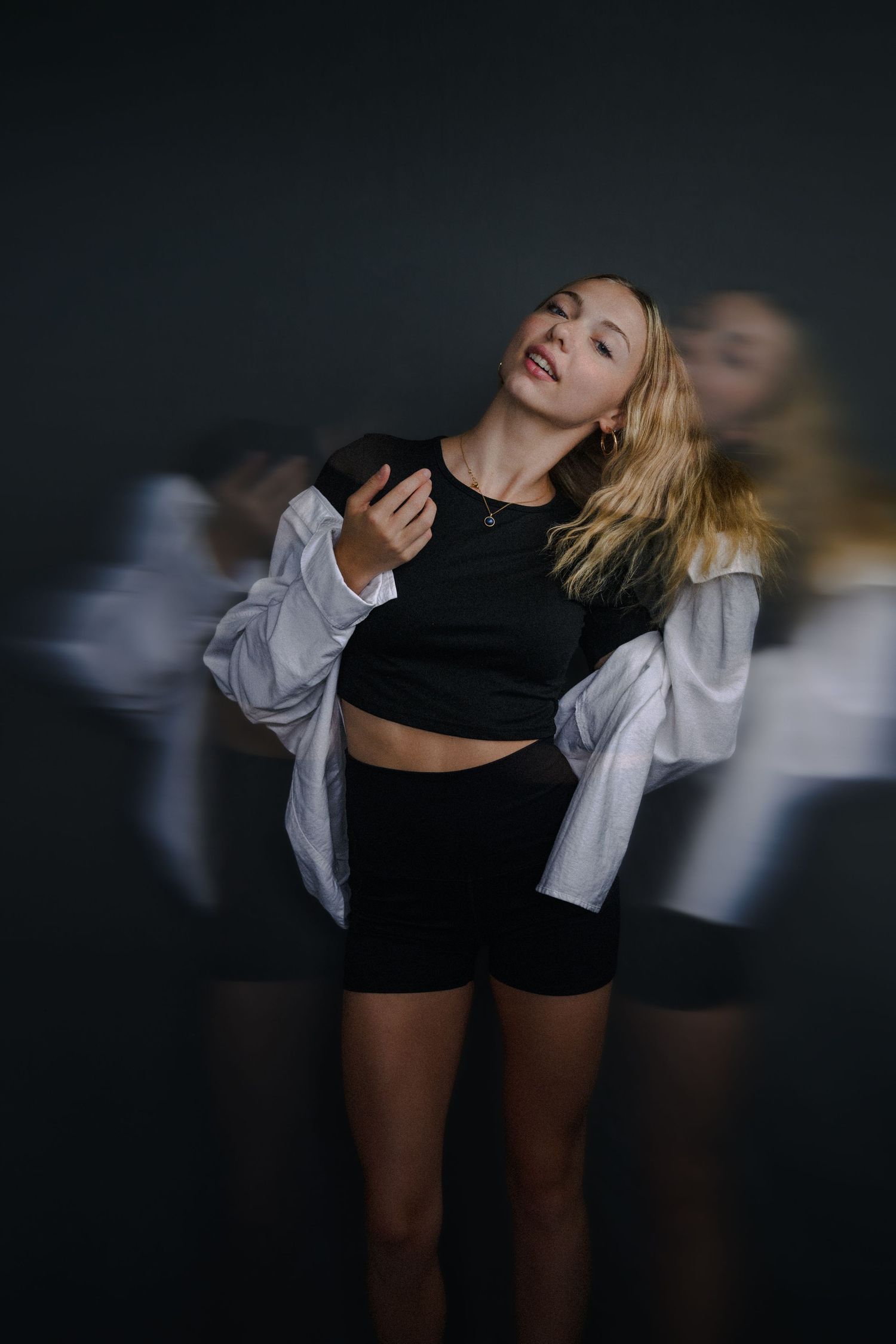 Blonde woman in black crop top and shorts with white shirt, posing dynamically with motion blur effect on dark background.